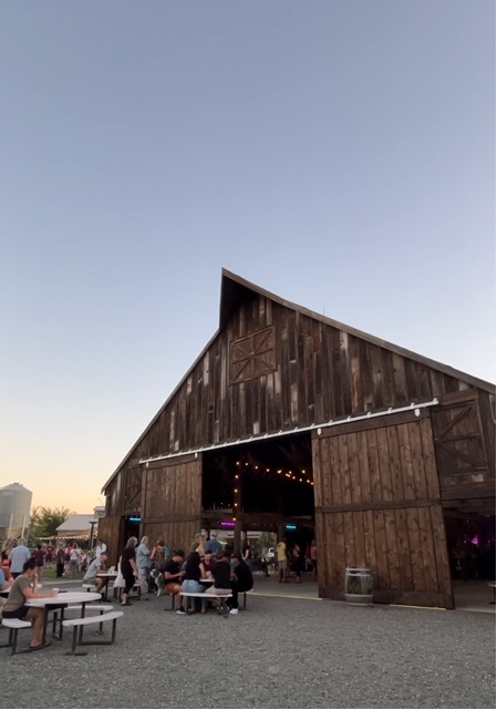 People gathered outside of a large wooden barn-style building during sunset, with some seated at picnic tables and others near the entrance with string lights overhead.