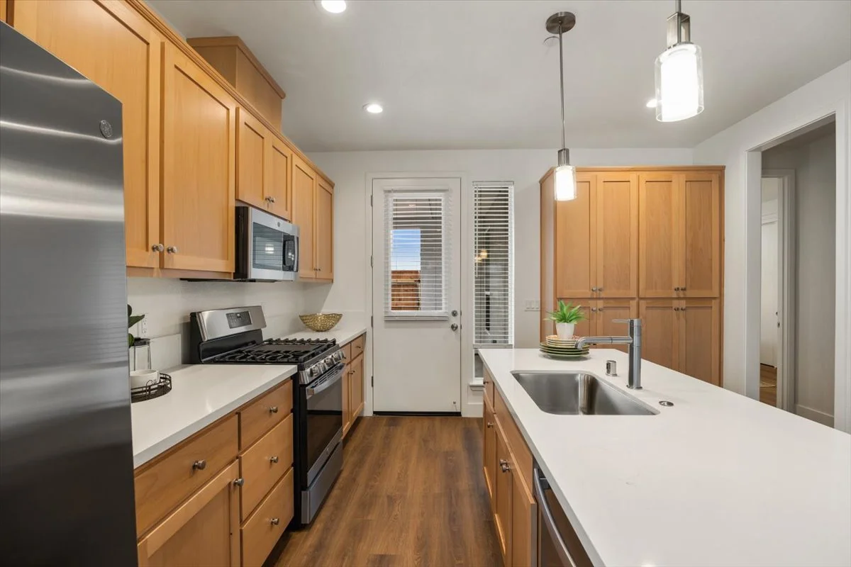 Modern kitchen with wooden cabinets, white countertops, stainless steel appliances, a sink on the island, and a door with blinds leading outside.