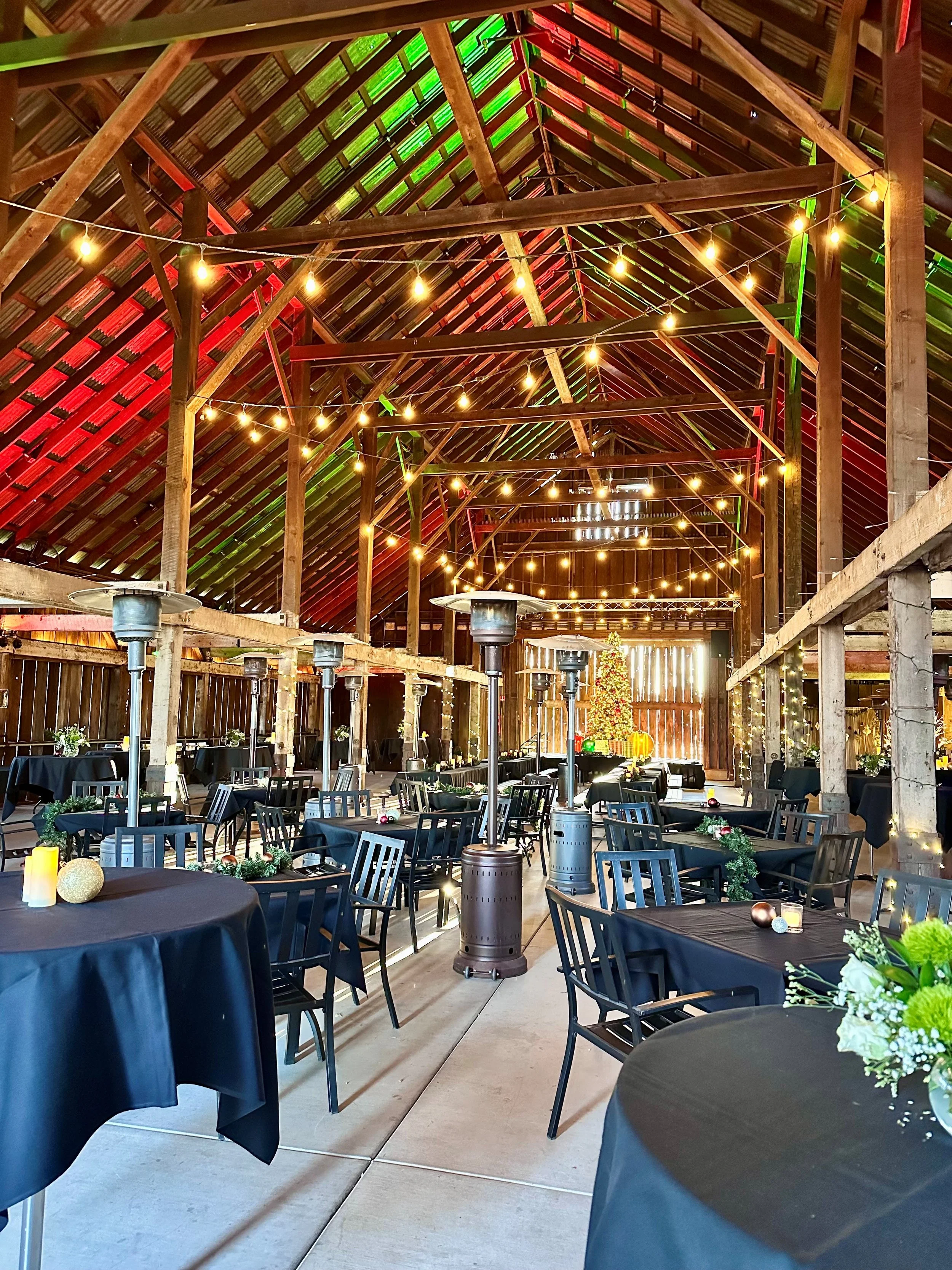 A decorated indoor event space with string lights, black-covered tables, and chairs, decorated with holiday ornaments and flowers, with a Christmas tree at the back.
