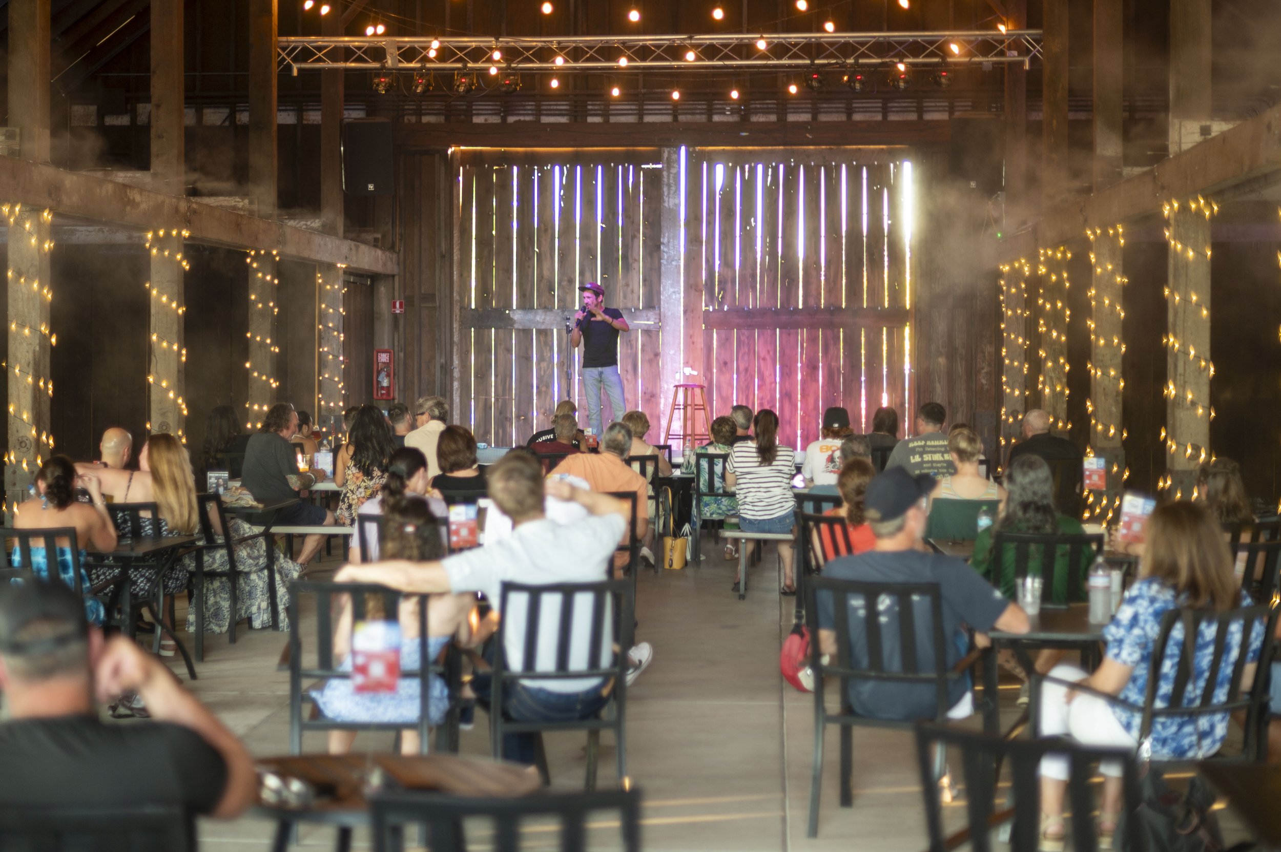 A comedy show or stand-up performance is taking place in a rustic venue with wooden walls and string lights. A comedian stands on a stage with a microphone, while an audience seated at tables watch the performance.