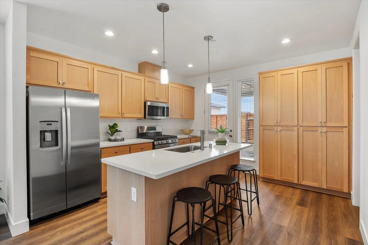 Modern kitchen with wooden cabinets, stainless steel refrigerator, microwave, gas stove, white countertop island with three black stools, and hardwood flooring.