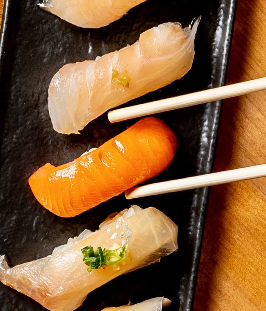Assorted sushi on a black rectangular plate, including slices of fish and a piece of salmon, with chopsticks resting on the plate.
