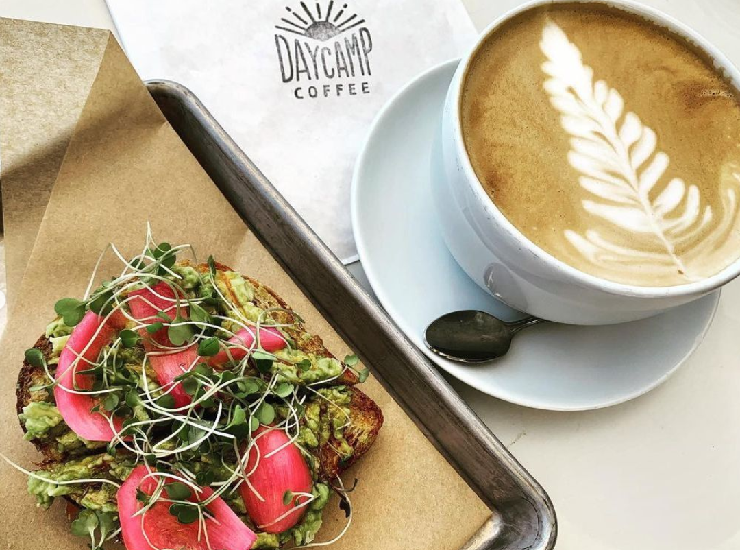 A cup of latte with latte art on top, a toasted bread topped with guacamole, radish slices, and microgreens, on a tray at Daycamp Coffee.