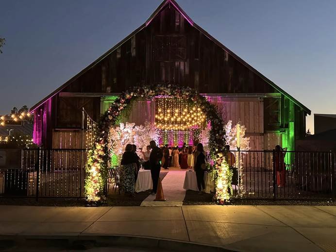 A barn decorated with pink and green lighting at night, featuring a flower arch with lights and guests inside for a celebration or event.