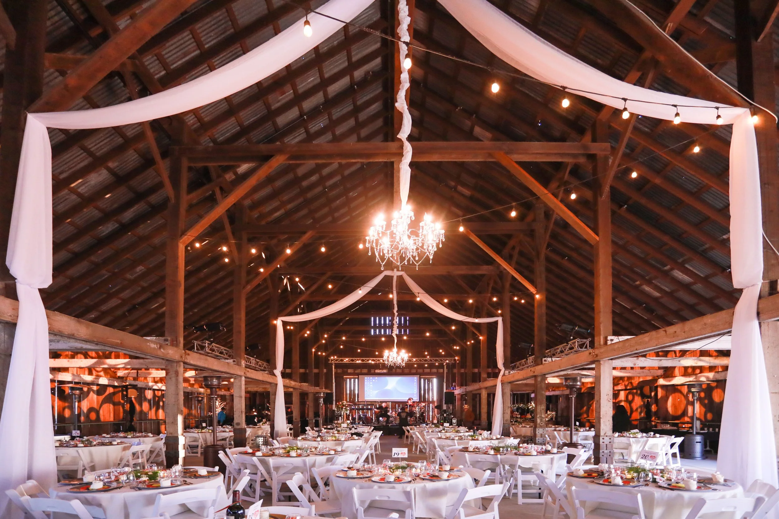 Wedding reception setup inside a wooden barn with round tables covered in white tablecloths, decorated with plates, glasses, and centerpieces. Elegant chandeliers and string lights are hanging from the ceiling with white drapes draped along the beams