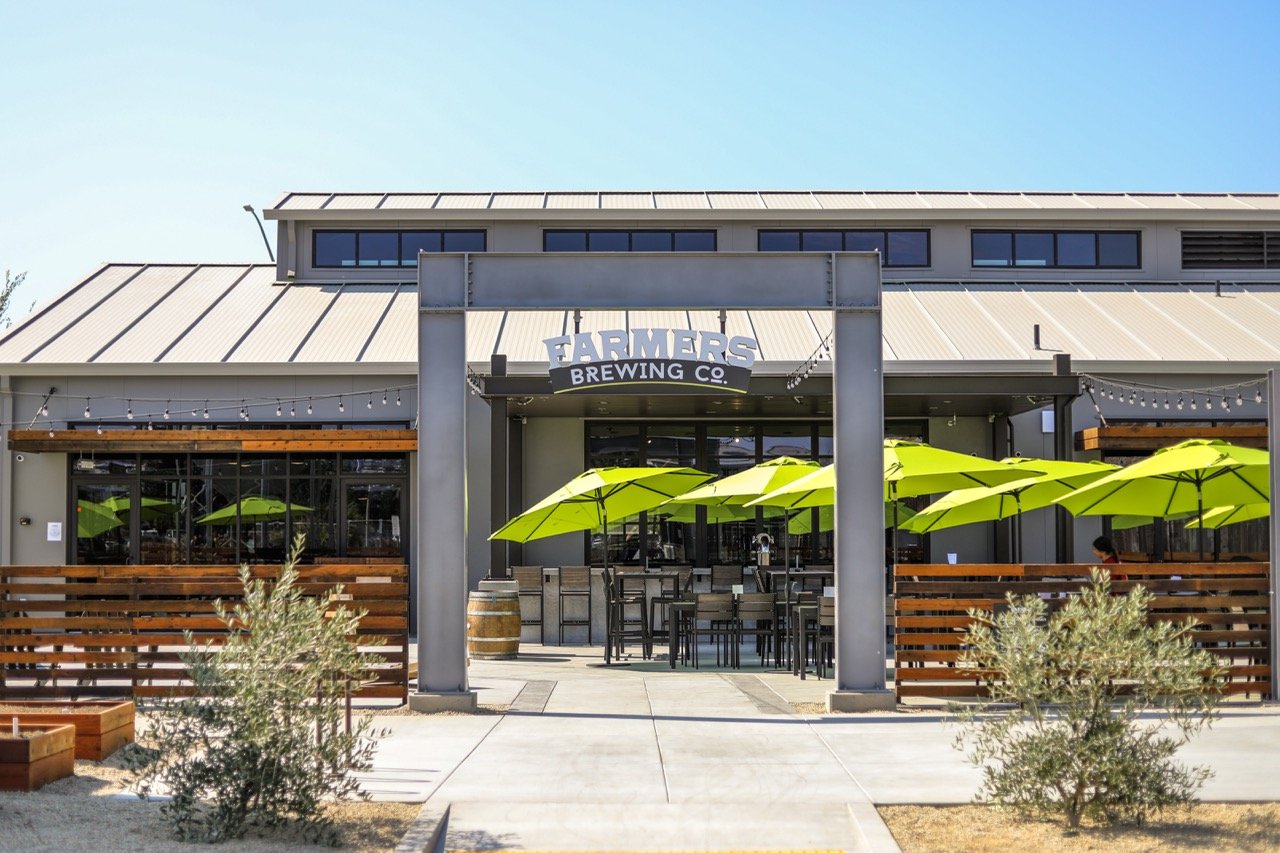 Exterior of Farmers Brewing Co. with outdoor seating, lime green umbrellas, and string lights under a clear blue sky.