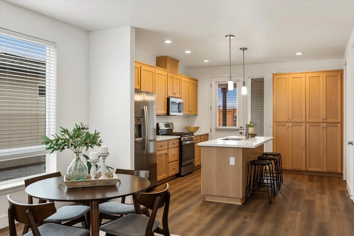 Modern kitchen with wooden cabinets, stainless steel appliances, a kitchen island with a white countertop, bar stools, a round dining table with chairs, and decorative elements like a vase with greenery and candles.