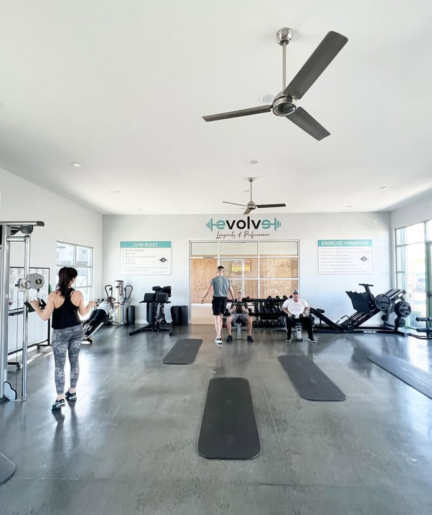 A gym with people working out and sitting on benches, with gym equipment, mats, and large windows allowing natural light.