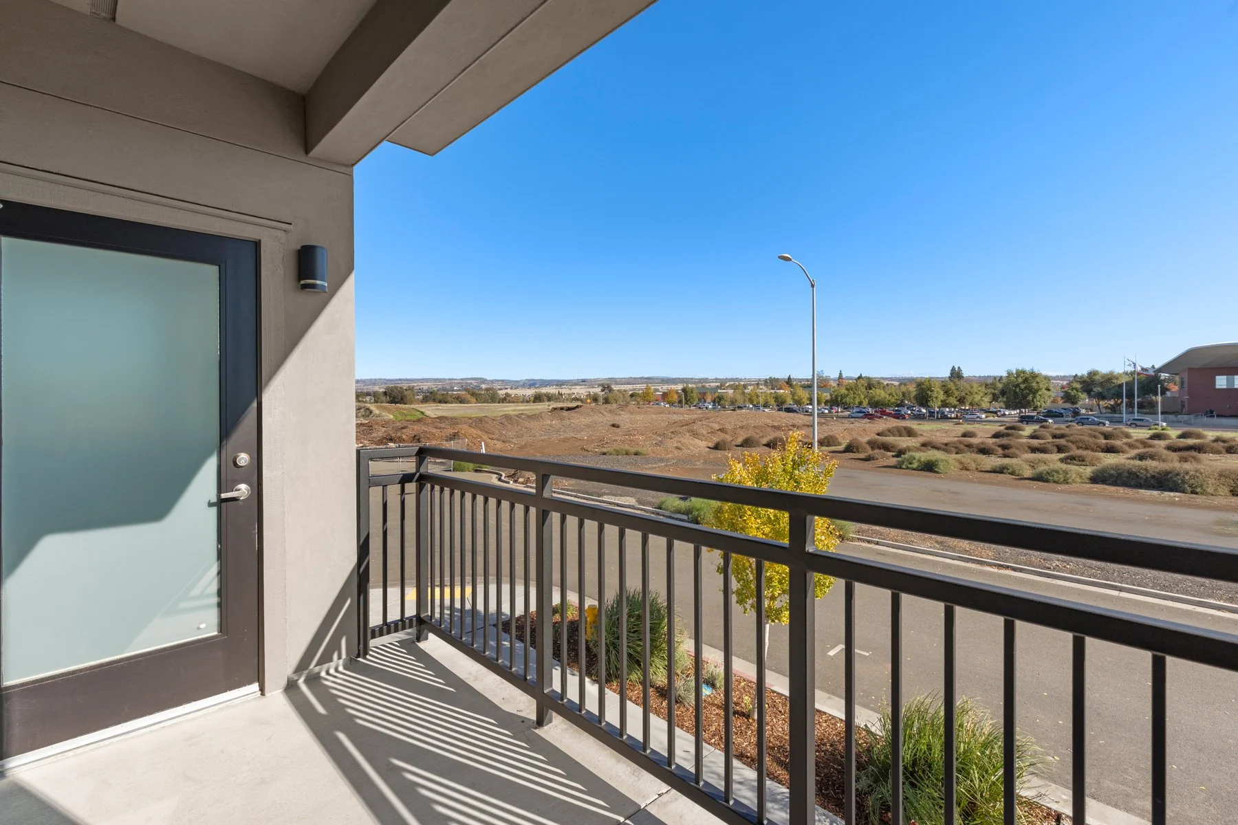 View from an apartment balcony showing a parking lot, a street lamp, and a distant landscape with trees and buildings under a clear blue sky.