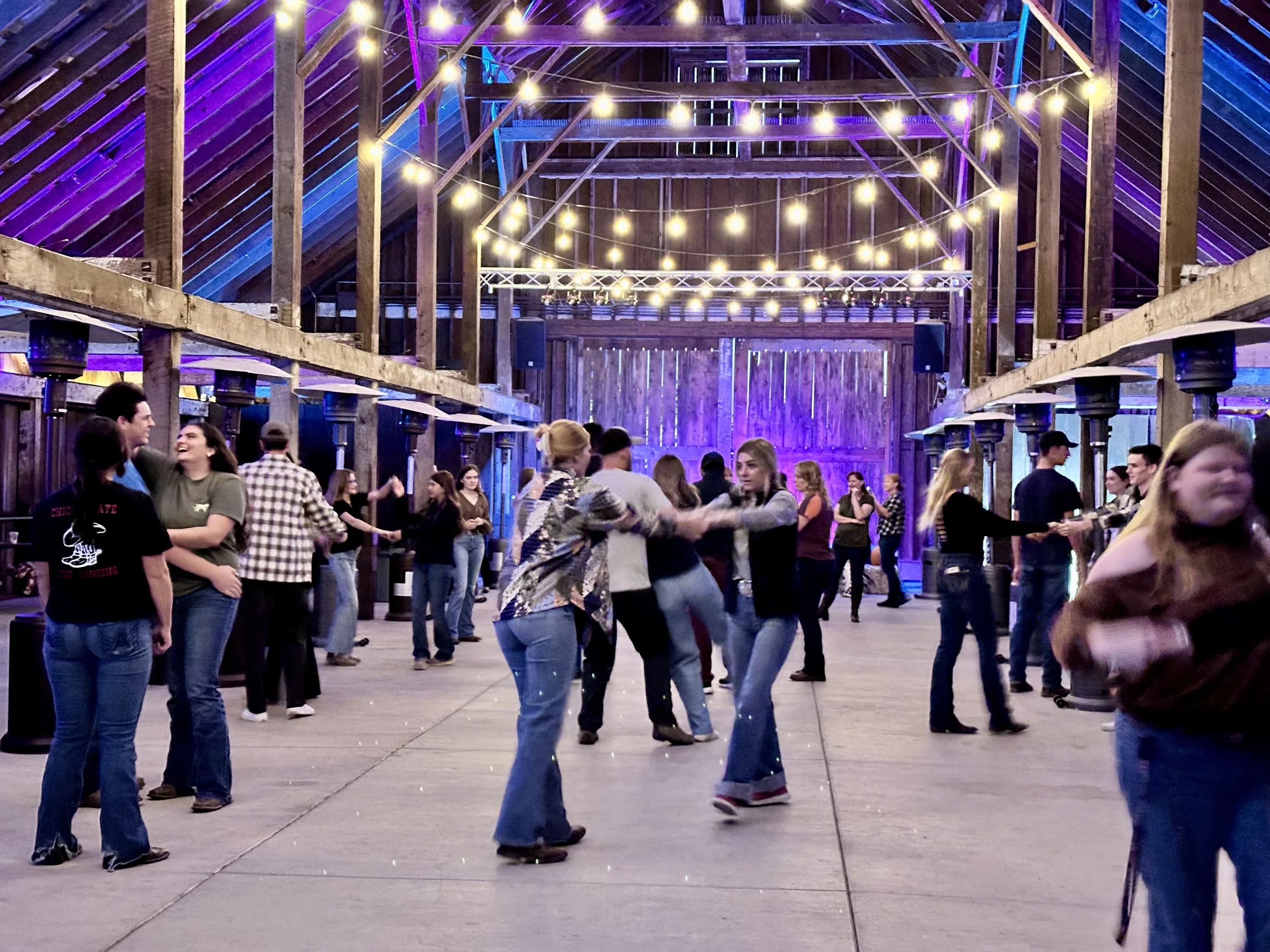 People dancing and socializing at a rustic barn party with string lights overhead and purple lighting on wooden walls.