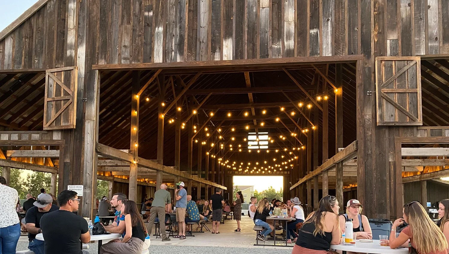 People socializing and dining in a rustic wooden barn with string lights hanging from the ceiling.