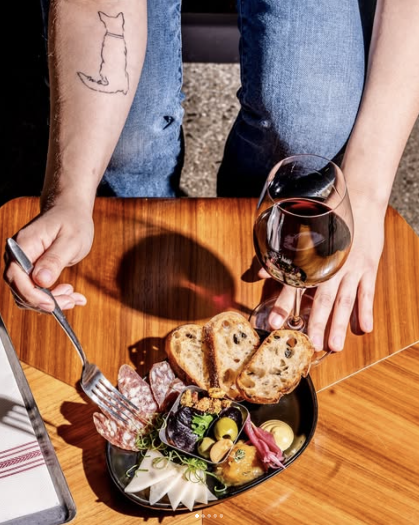 Person holding a glass of red wine and a plate of assorted charcuterie, bread slices, and snacks on a wooden table.