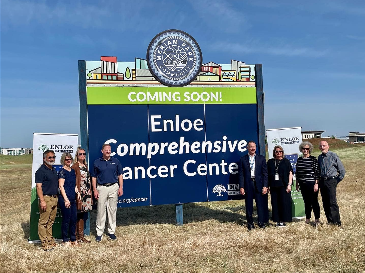 Group of seven people standing next to a large sign that reads 'Coming Soon! Enloe Comprehensive Cancer Center' with the Enloe Medical Center logo, outdoors in a grassy area under a blue sky.
