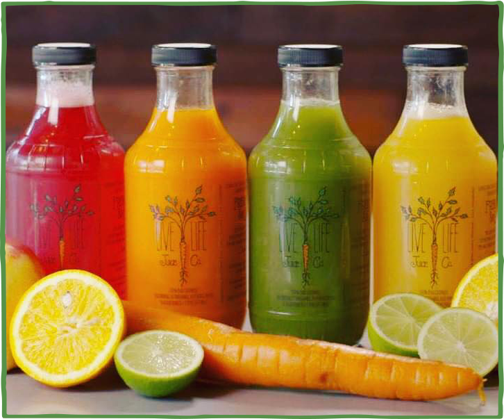Four bottles of colorful fruit juice lined up, with fresh lemons, limes, and a carrot in front of them.