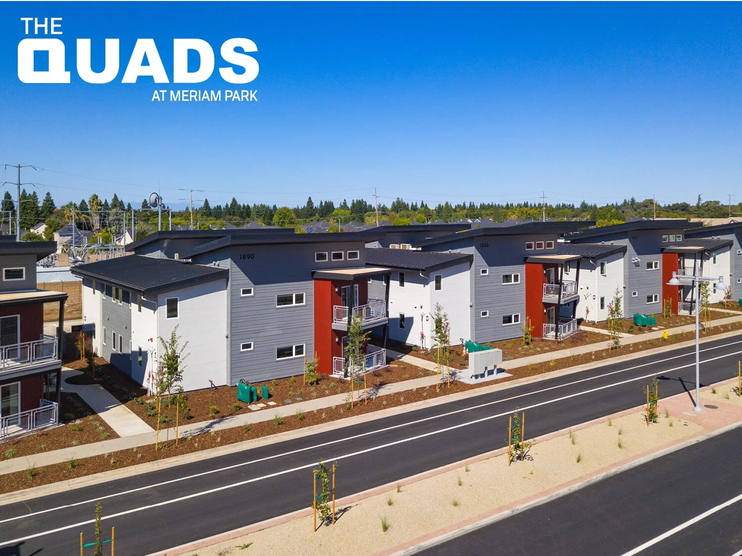 Aerial view of newly constructed modern multi-family housing units at Meriam Park, with neatly landscaped sidewalks, young trees, and an empty street under a clear blue sky.