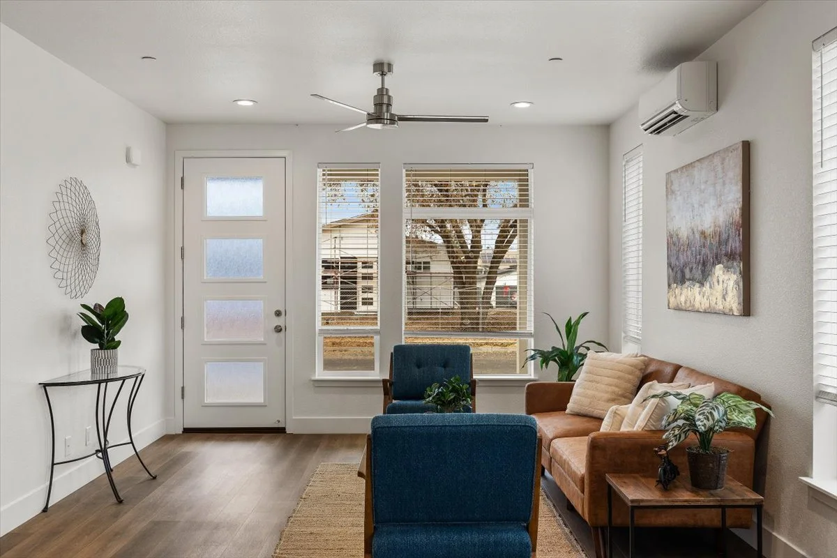 Living room with white walls, hardwood floors, a front door with three glass panels, large windows with blinds, a ceiling fan, an air conditioning unit, a brown sofa with pillows, two blue armchairs, a small side table with a plant, and wall art.