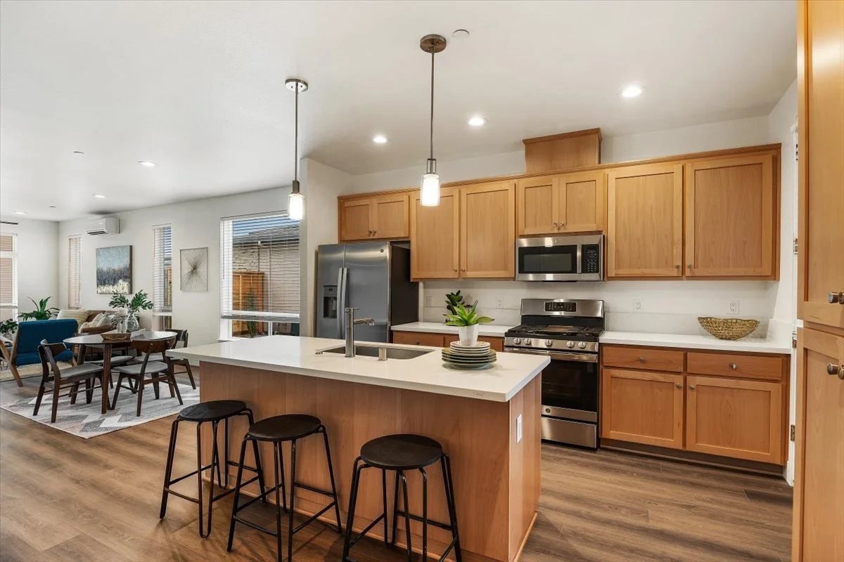 Open-concept kitchen with wooden cabinets, stainless steel appliances, white countertops, and a kitchen island with three black stools. In the background, there is a living and dining area with large windows, wooden furniture, and decor.