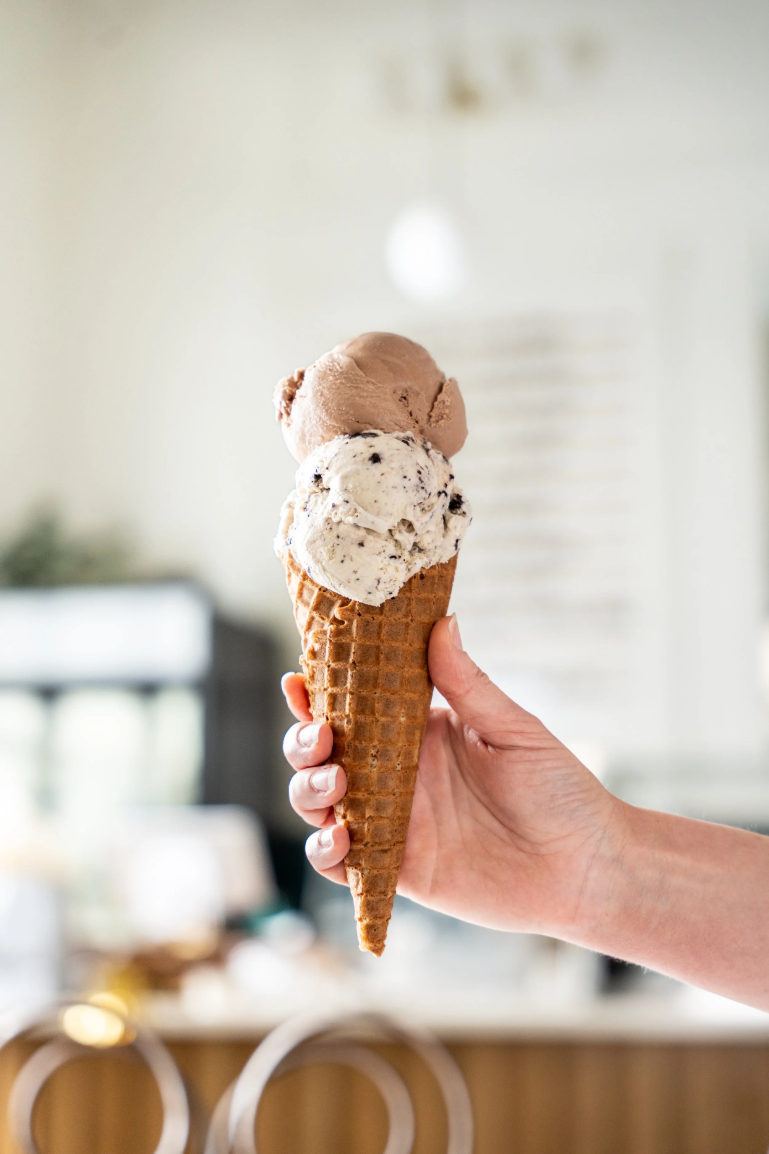 A hand holding an ice cream cone with two scoops of ice cream, one with chocolate chips and the other plain, inside a bright, modern kitchen.