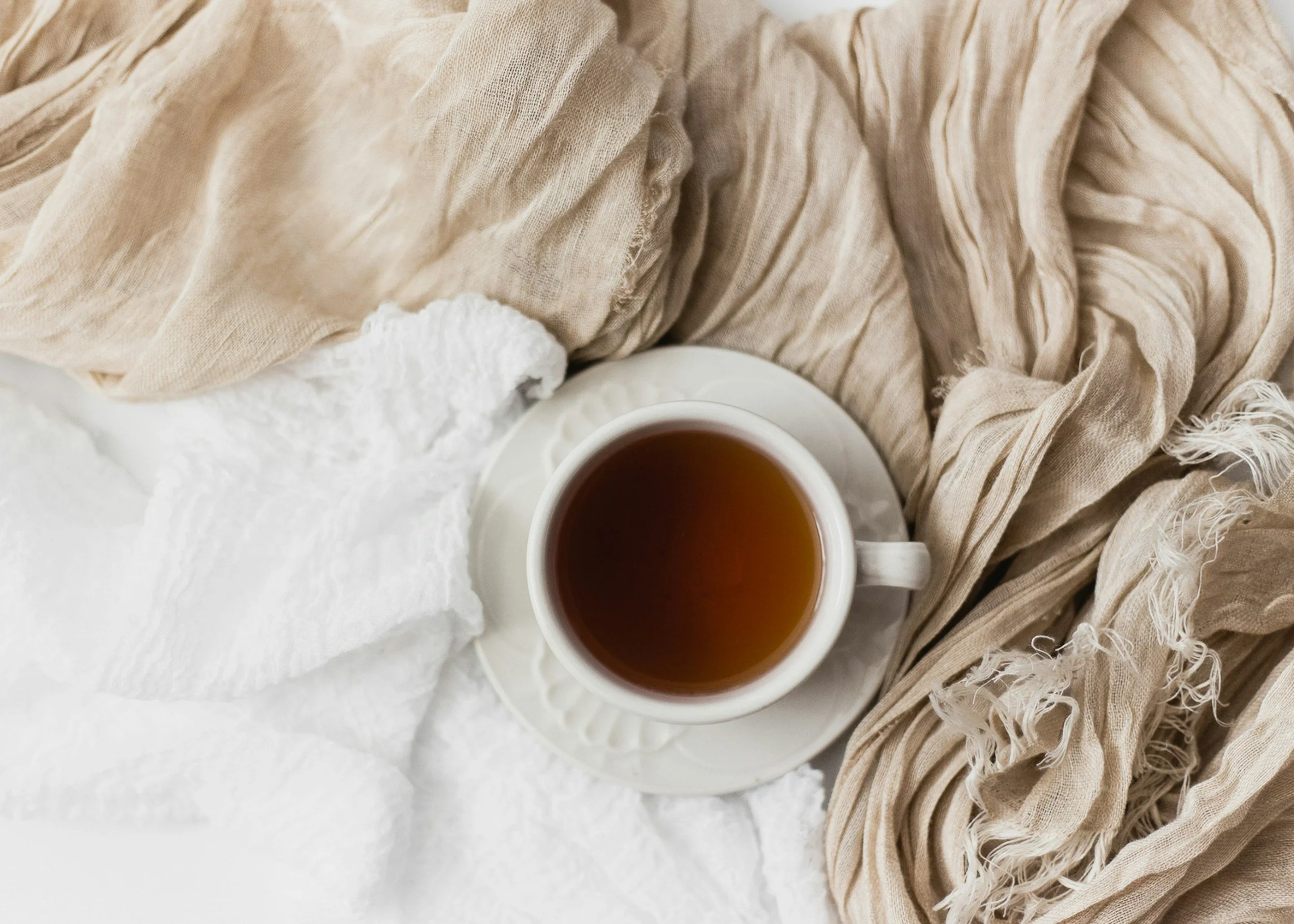 A white cup of tea resting on a saucer, surrounded by crumpled beige fabric and a piece of white cloth, on a white surface.