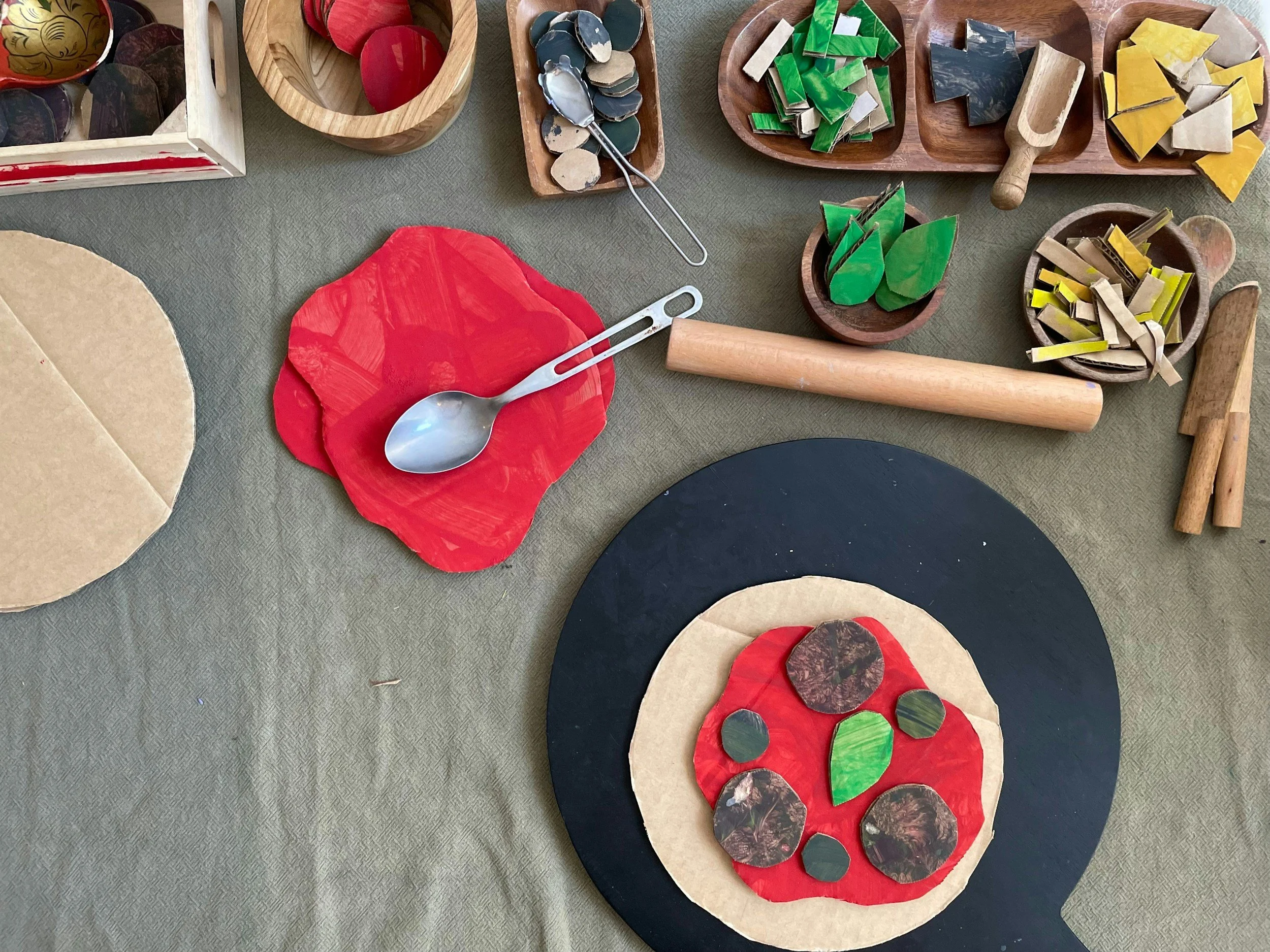 Child's art project with paper cutouts resembling a pizza with red, green, and brown toppings, along with various craft utensils like spoons, rolling pin, and small wooden bowls with colored paper pieces for creating a pizza design.