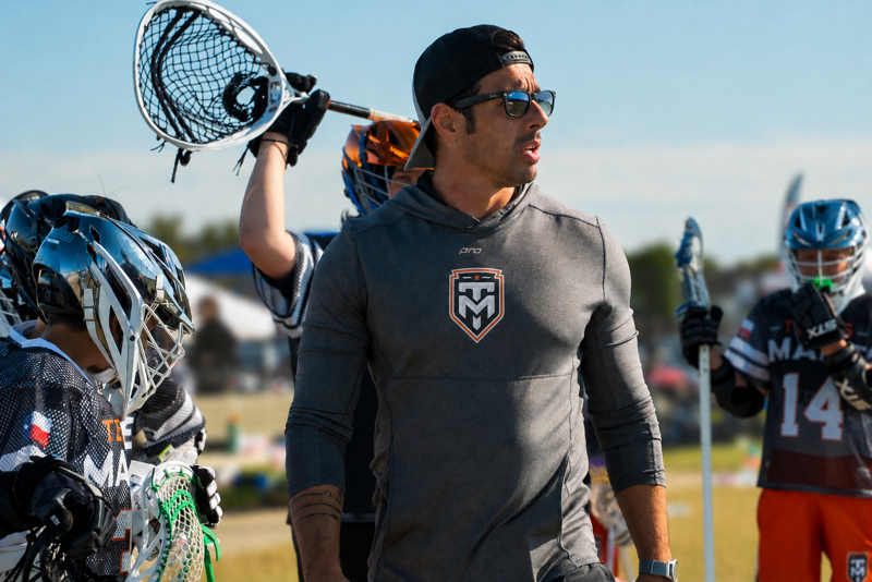 Lacrosse coach stands on the sideline with youth players holding lacrosse sticks during an outdoor Texas Made Lacrosse event.
