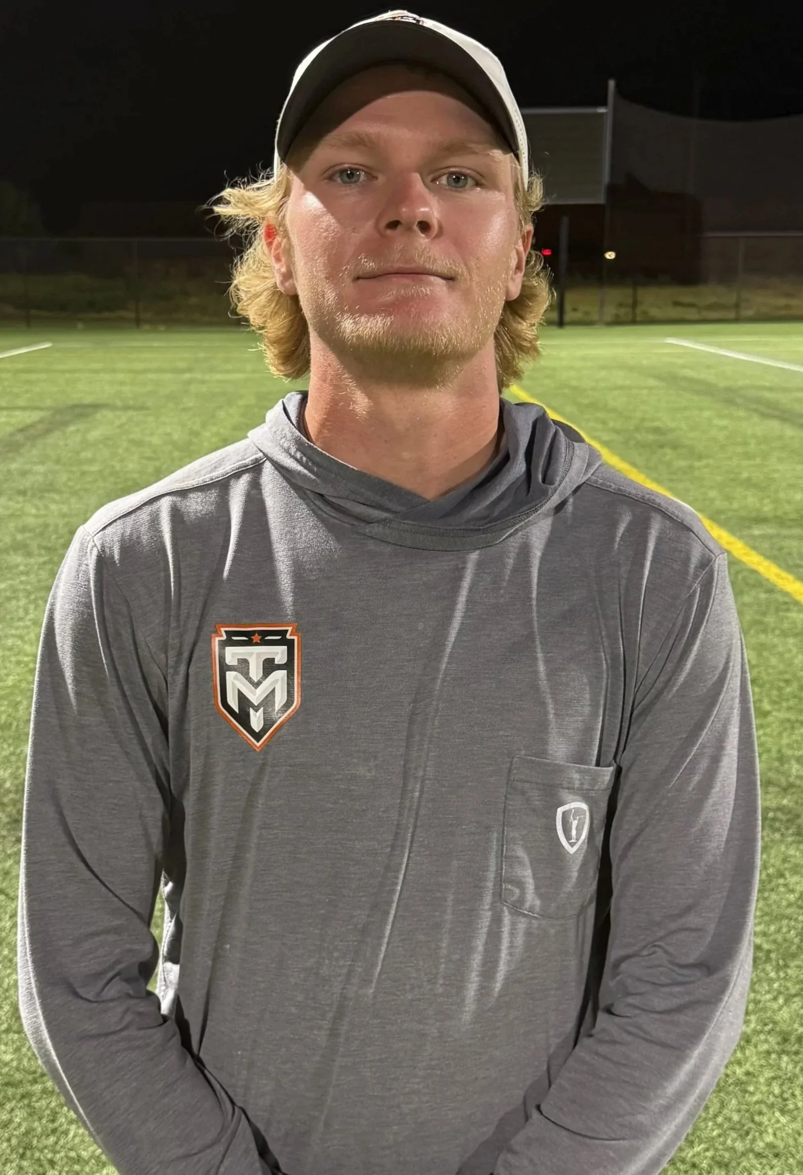 Jonah Heaton wearing a gray Texas Made lacrosse hoodie and hat on the field at night.