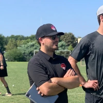 Cory Childs in a black polo with sunglasses his arms crossed and a iPad tucked under his arm on a lacrosse field.