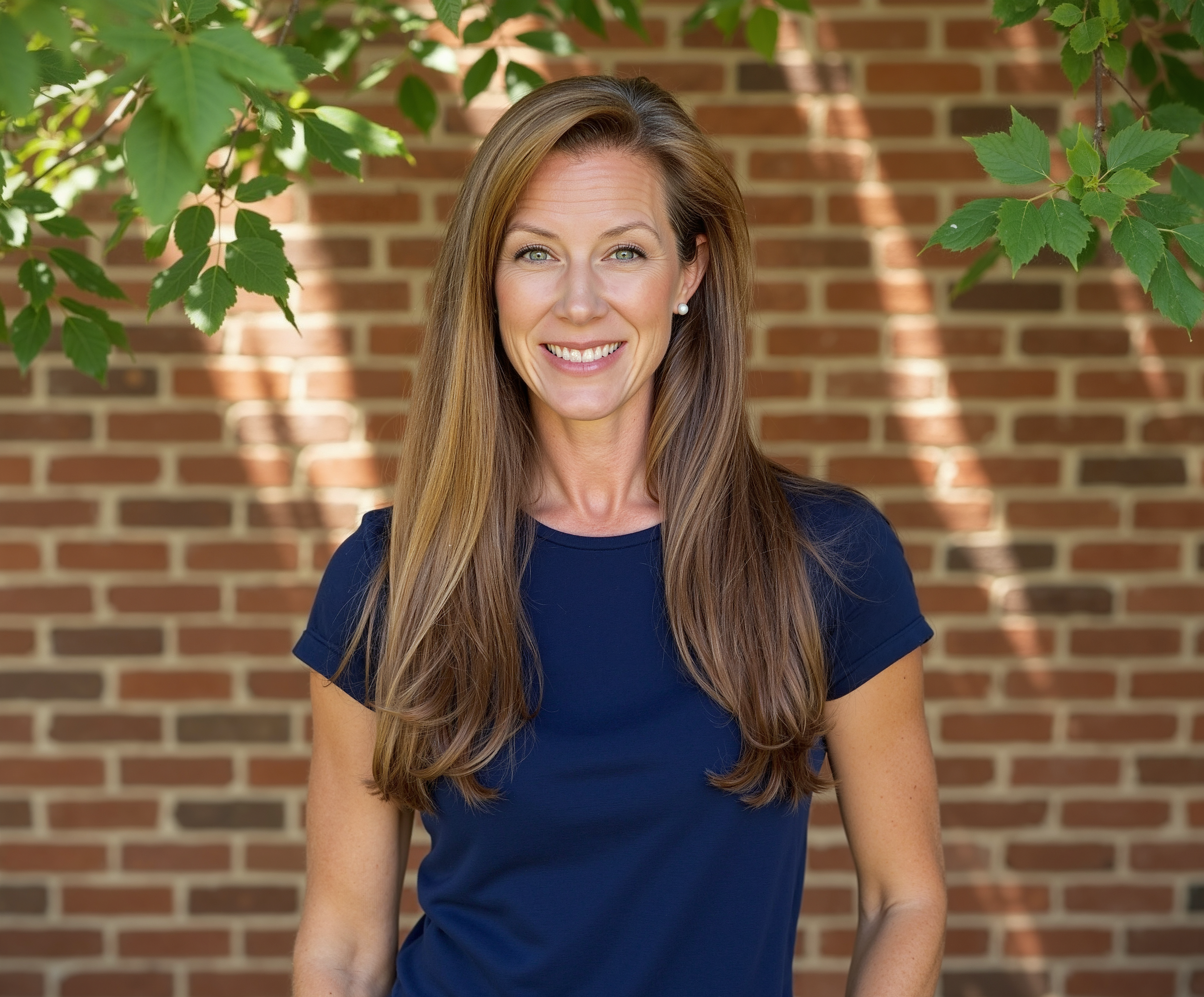 A picture of Whitney Ashworth Kopco with long brown hair, wearing a navy blue shirt, smiling outdoors in front of a brick wall and green leafy branches.