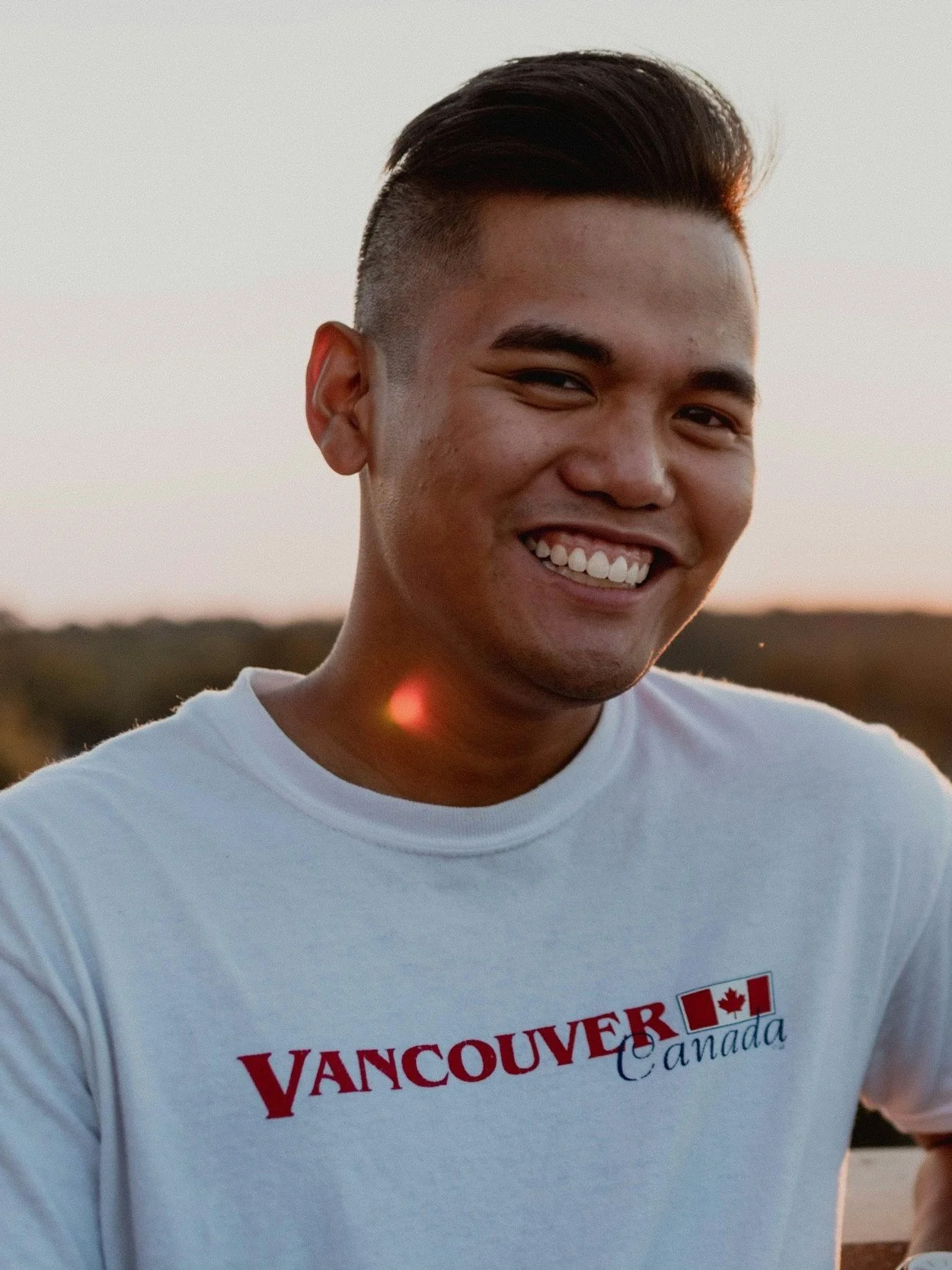 A smiling young man with short dark hair, wearing a white Vancouver, Canada t-shirt, standing outdoors during sunset.