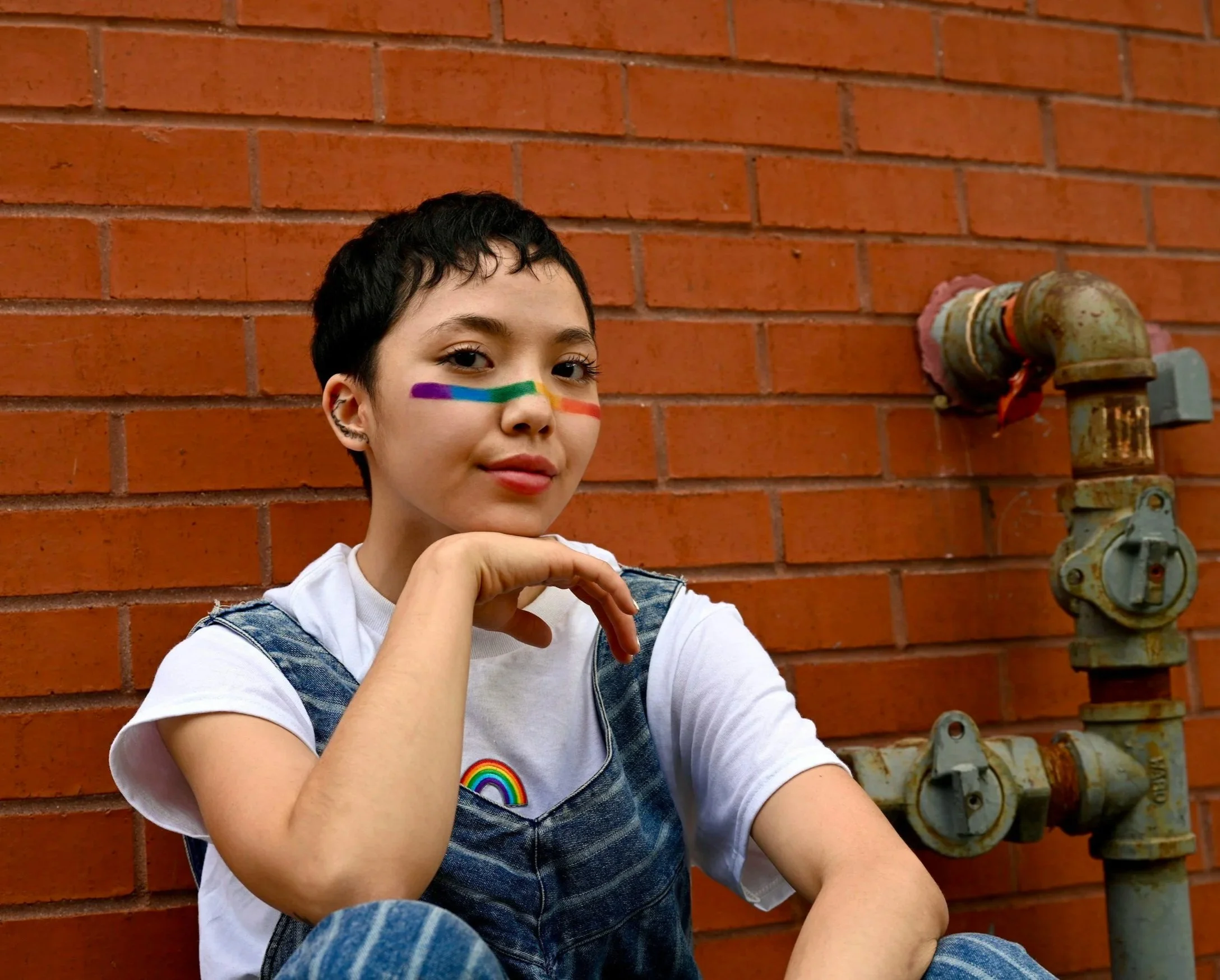 A young person with short dark hair and rainbow face paint on their cheek, sitting against a red brick wall with a rusty water pipe and valve.