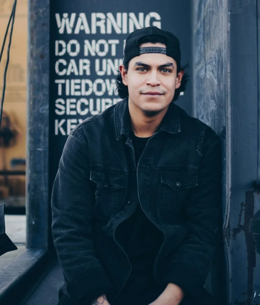 A young man with dark hair, wearing a black backwards cap and black denim jacket, sitting outside near a black wall with a warning sign in the background.