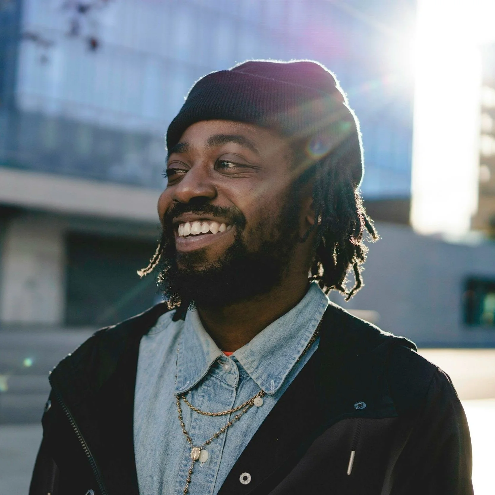 A smiling man with dreadlocks wearing a black beanie, layered necklaces, a light denim shirt, and a black jacket standing outdoors with sunlight shining behind him.