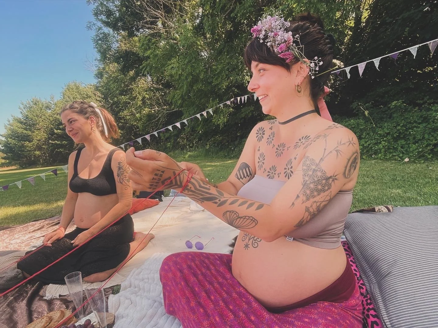 Two women with tattoos sitting outdoors on blankets, one is knitting, and the other is smiling. They are surrounded by trees and grass under a sunny sky.