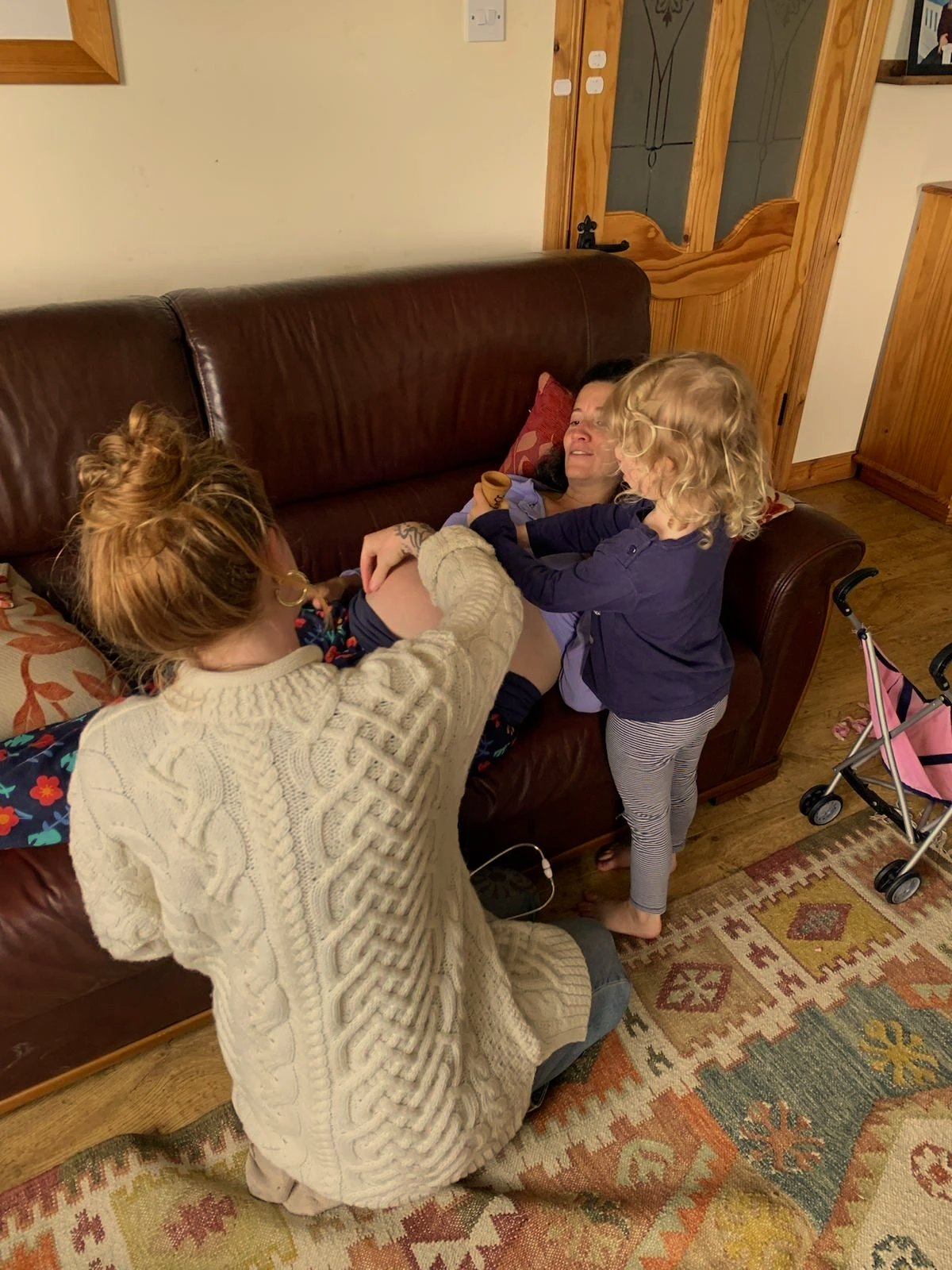 A woman is lying on a couch while two children, a girl with blond curly hair and a girl with red hair, are playfully tugging at her shirt in a cozy living room.