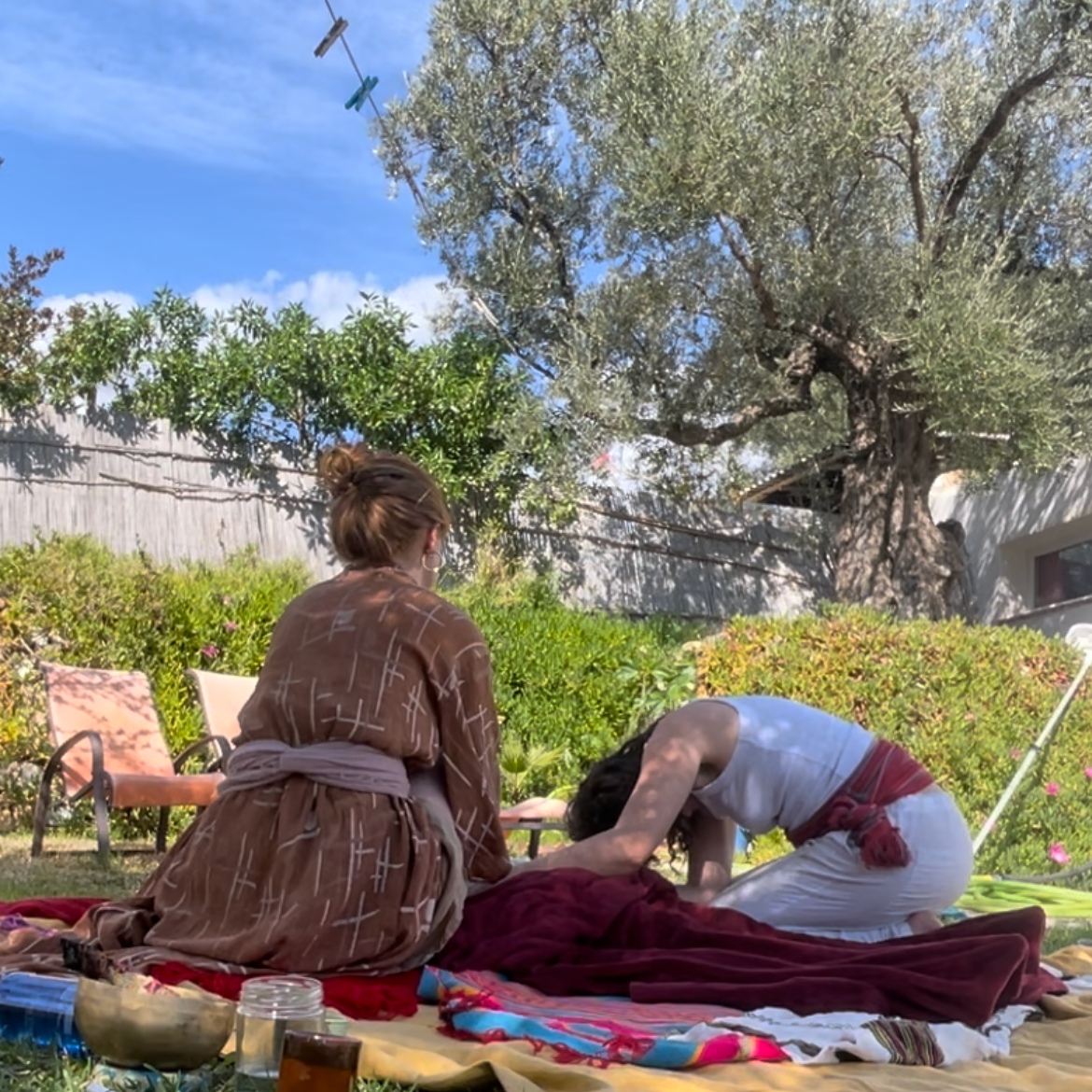 Two women practicing a traditional or spiritual ritual outdoors on a blanket in a garden with green bushes and a large tree.