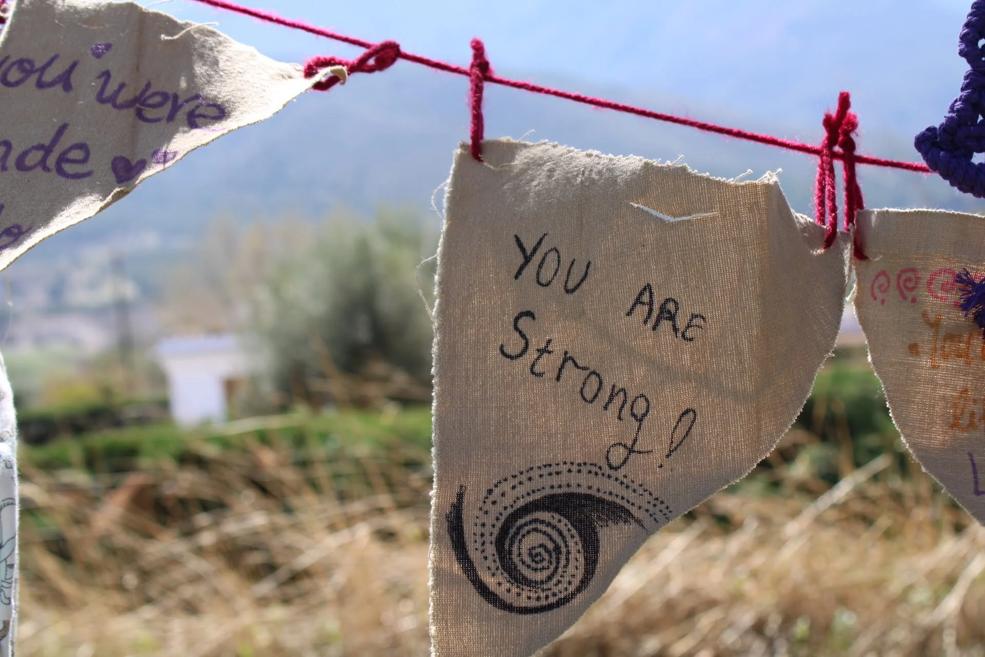 Fabric banners with handwritten notes hung on a pink string outdoors, one reading "You are strong!" with a spiral design, against a blurred natural landscape and cloudy sky.