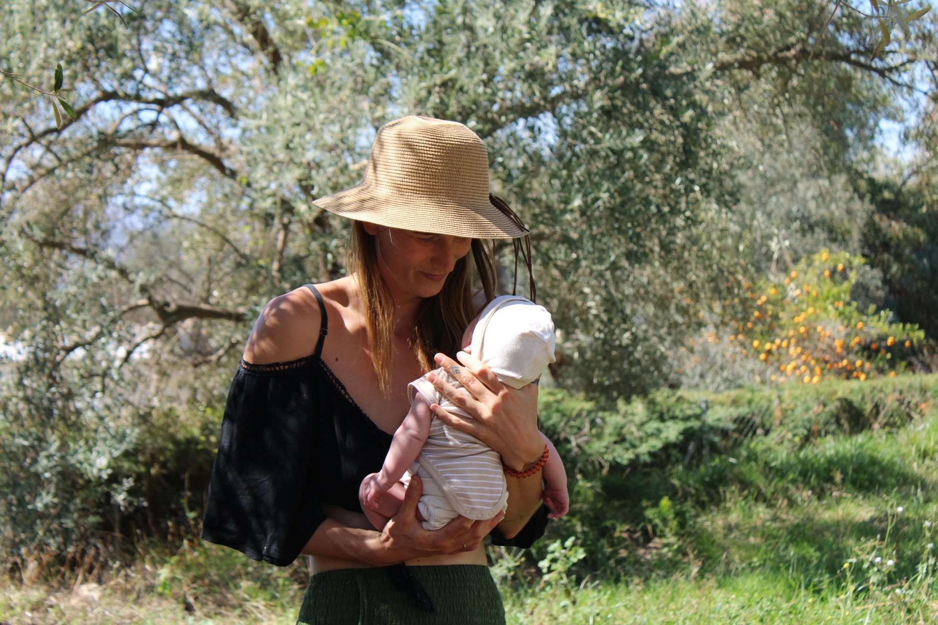 Woman wearing a straw hat holding a baby outdoors in a grassy area with trees and shrubs.