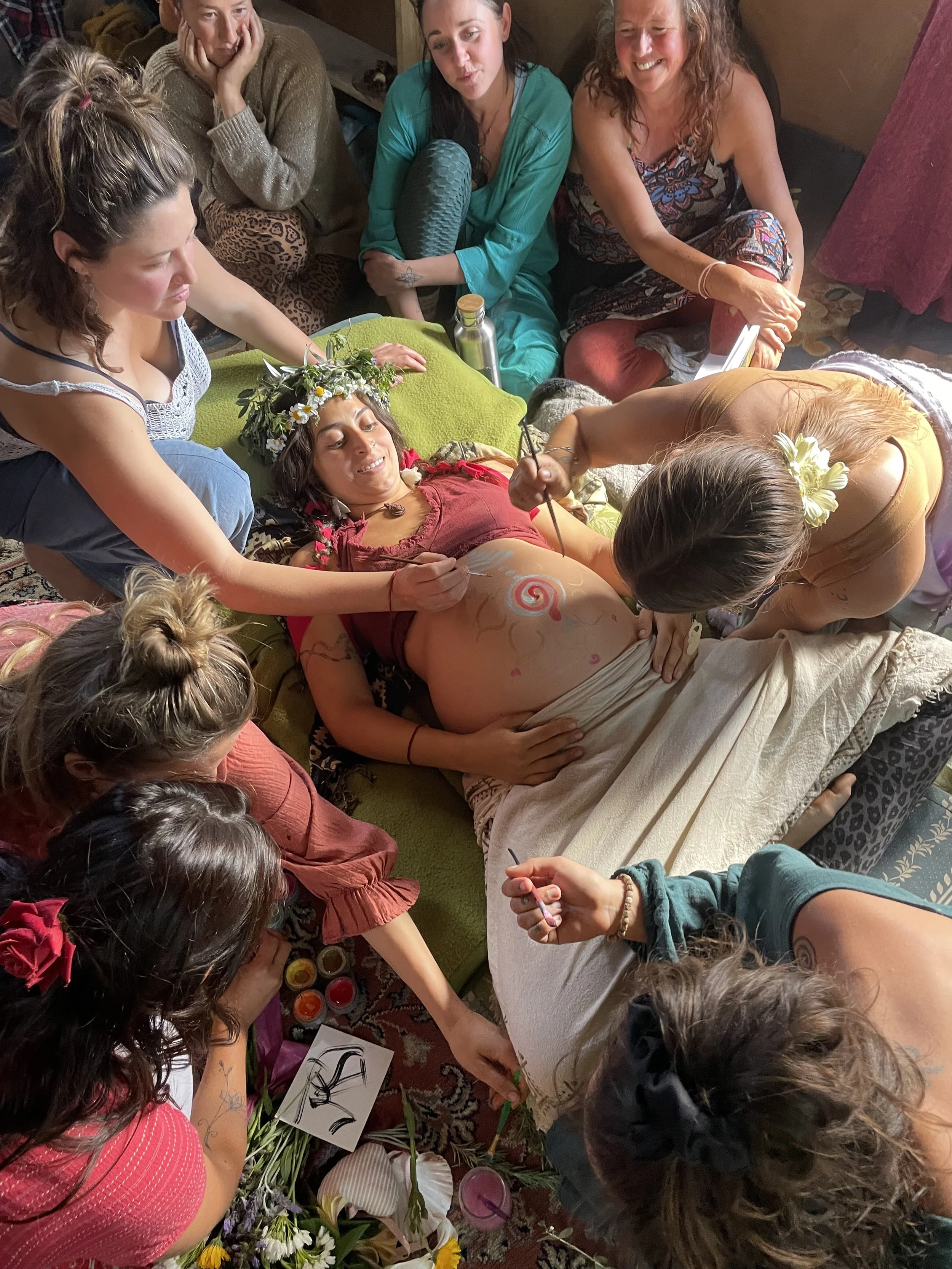 A woman lying on her back on a green sofa surrounded by friends, during a belly painting session. She wears a flower crown and a red shirt. Friends, both women and men, sit and kneel around her, painting a spiral on her belly, which is covered with various art supplies and flowers.