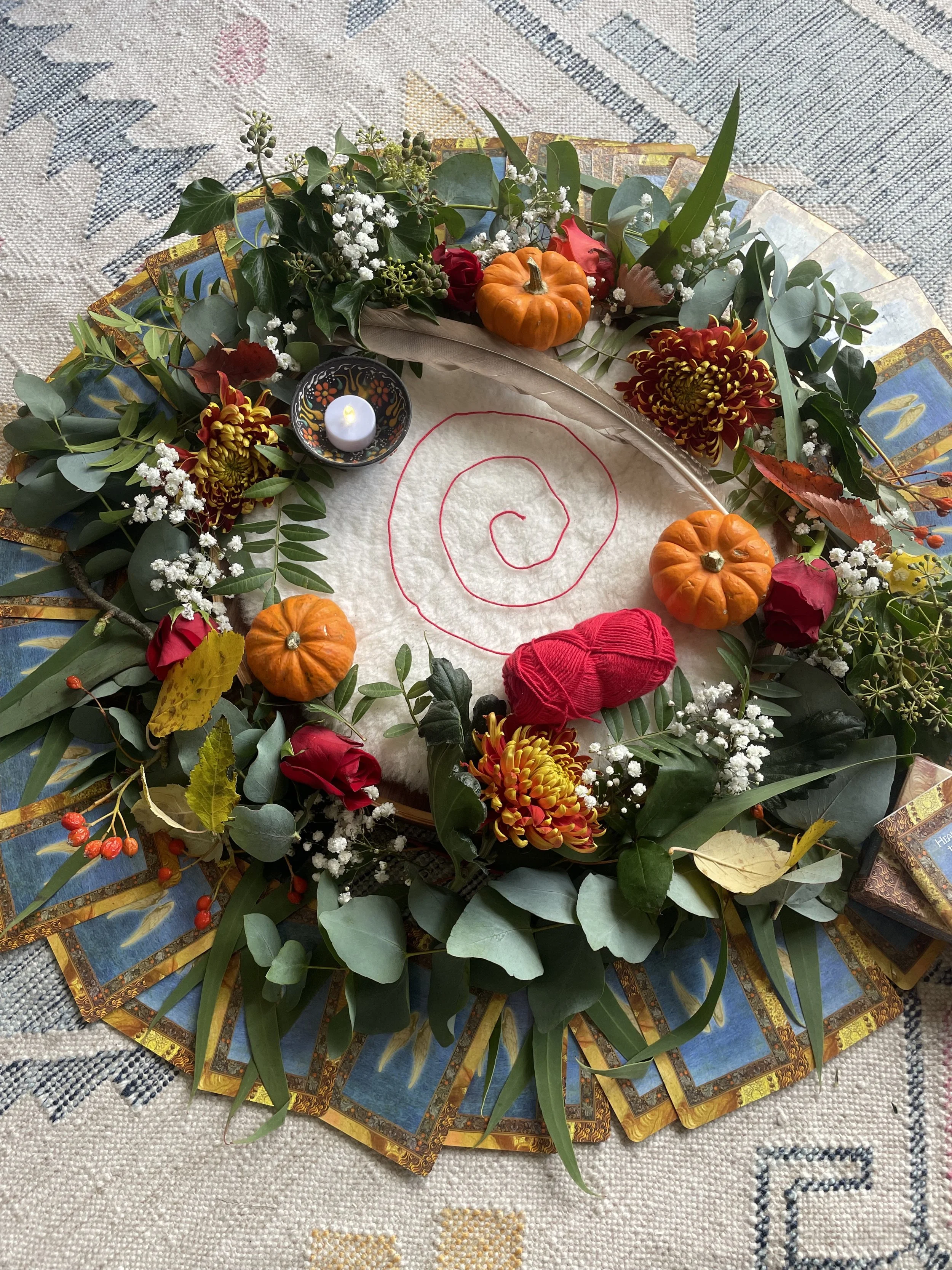 Autumn-themed centerpiece with small pumpkins, fall flowers, greenery, a lit tealight, tarot cards, and a red yarn ball, arranged on a decorative cloth.