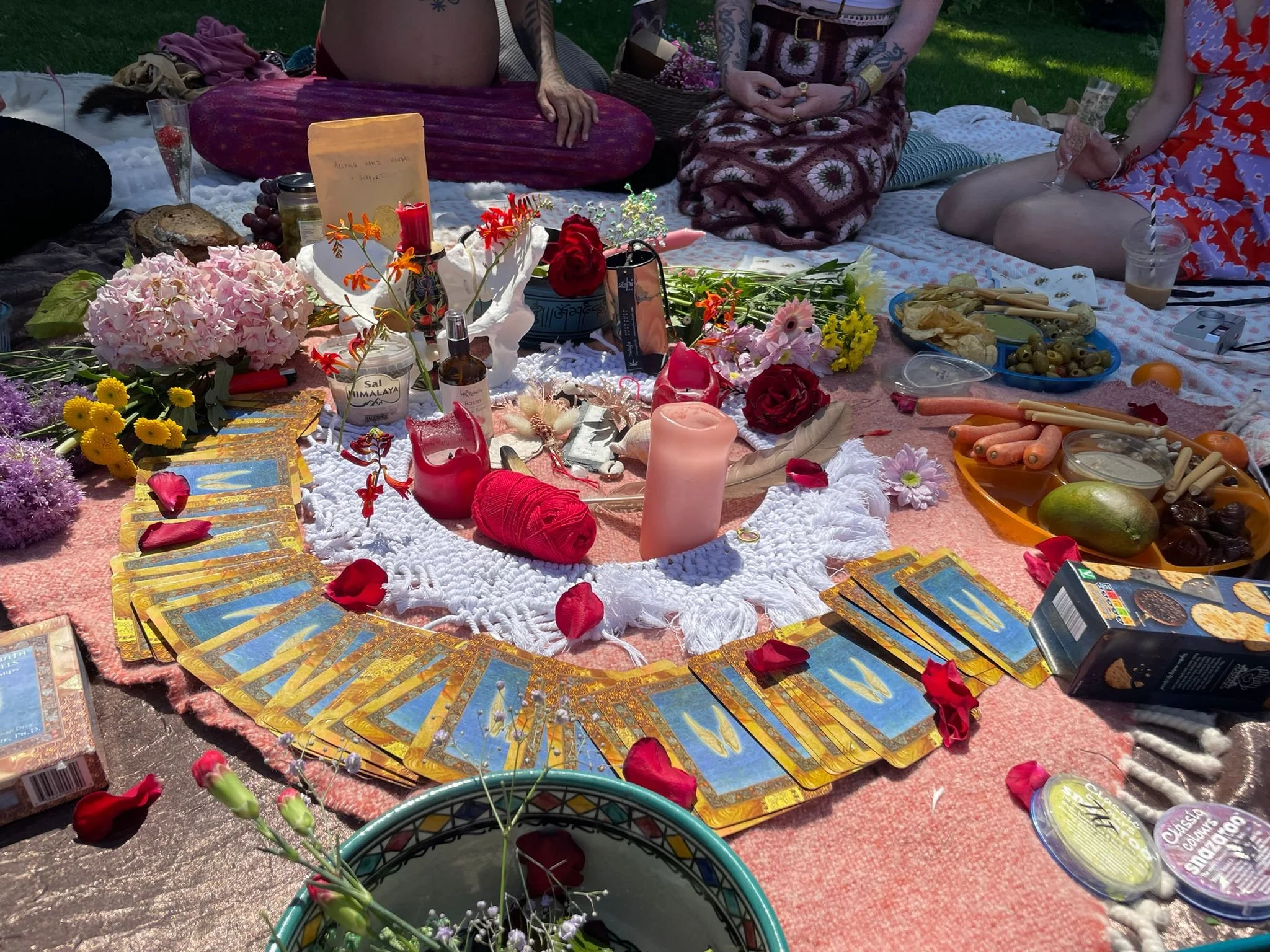 A colorful outdoor spiritual or ritual setup with tarot cards, candles, flowers, and various snacks on a blanket, surrounded by people sitting cross-legged.
