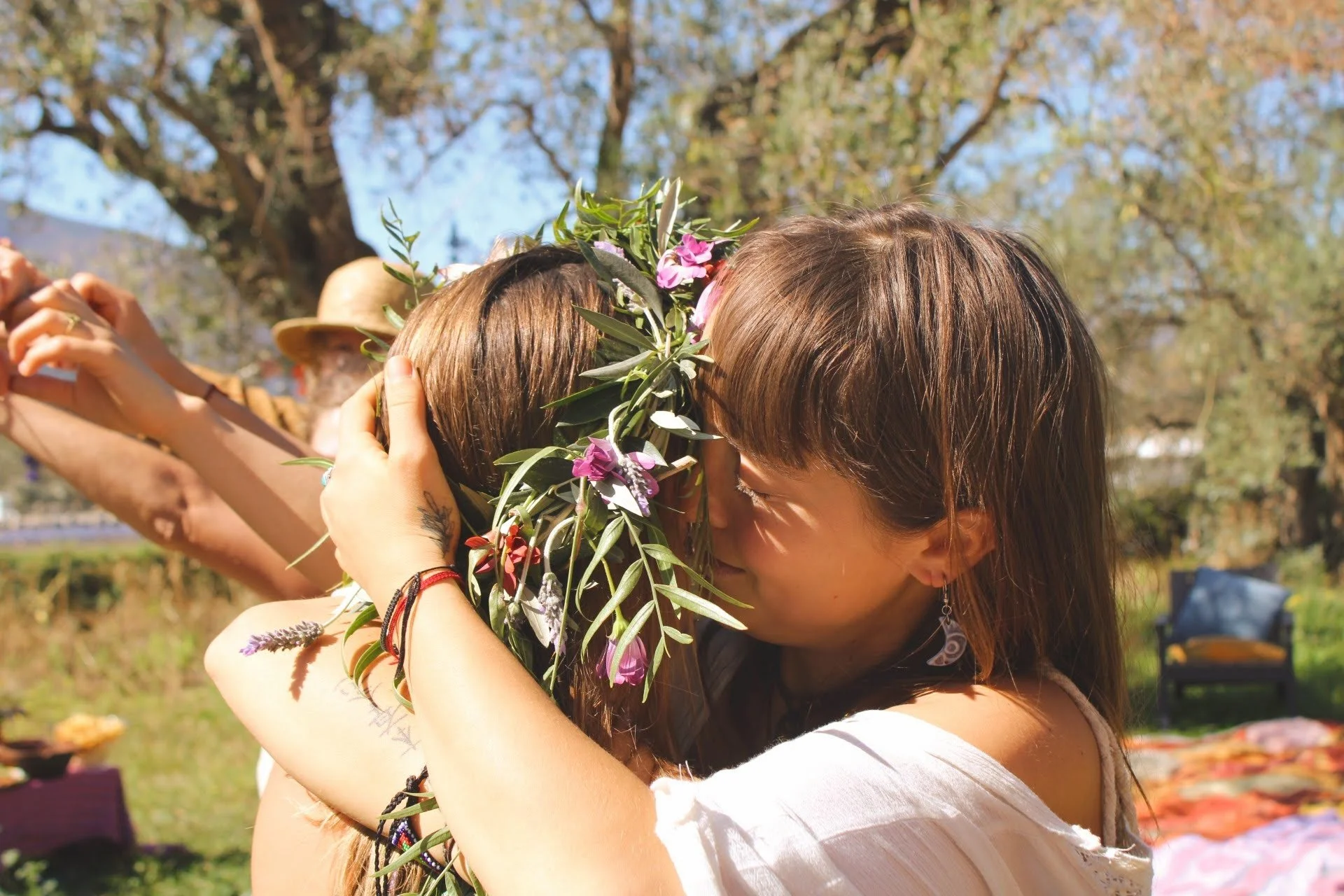 Two women hugging outdoors during daytime, one with a floral crown, with trees in the background.
