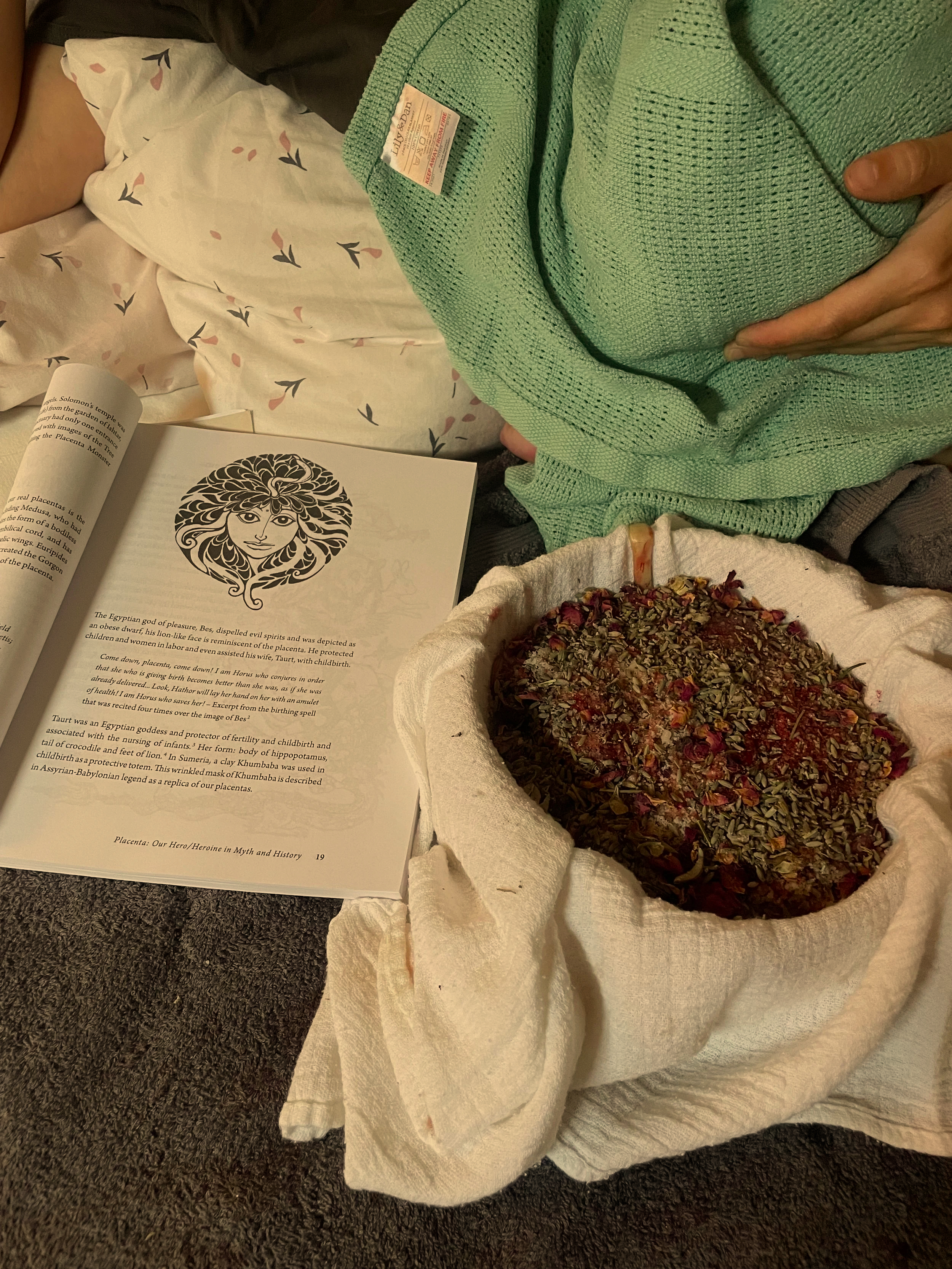 A person lying in a bed with white sheets decorated with black and pink leaves, holding a green knit blanket, next to an open book with an illustration of Medusa and text about Egyptian goddess Isis, and a round container filled with dried herbs and flowers, covered with a cloth.