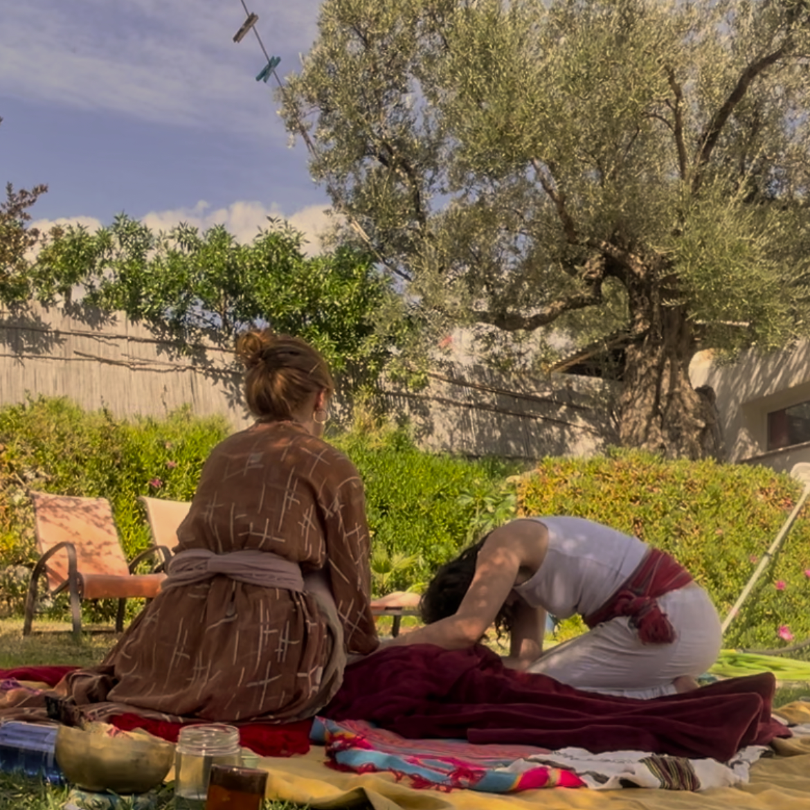 Two women practicing yoga or meditation outdoors on blankets in a backyard, surrounded by lush greenery, with a large tree and a wooden fence in the background.