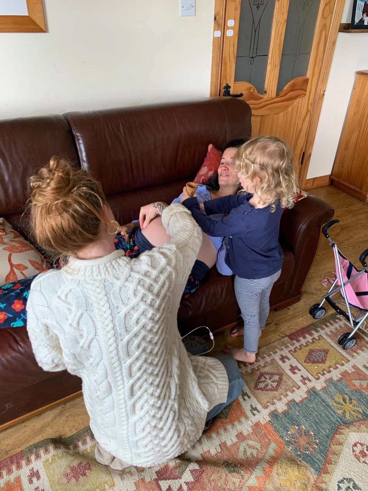 A woman lying on a brown leather couch while two young girls are playing with her, one of the girls is touching the woman's belly. All are in a cozy living room with a patterned rug, a pink stroller nearby, and wooden furniture.