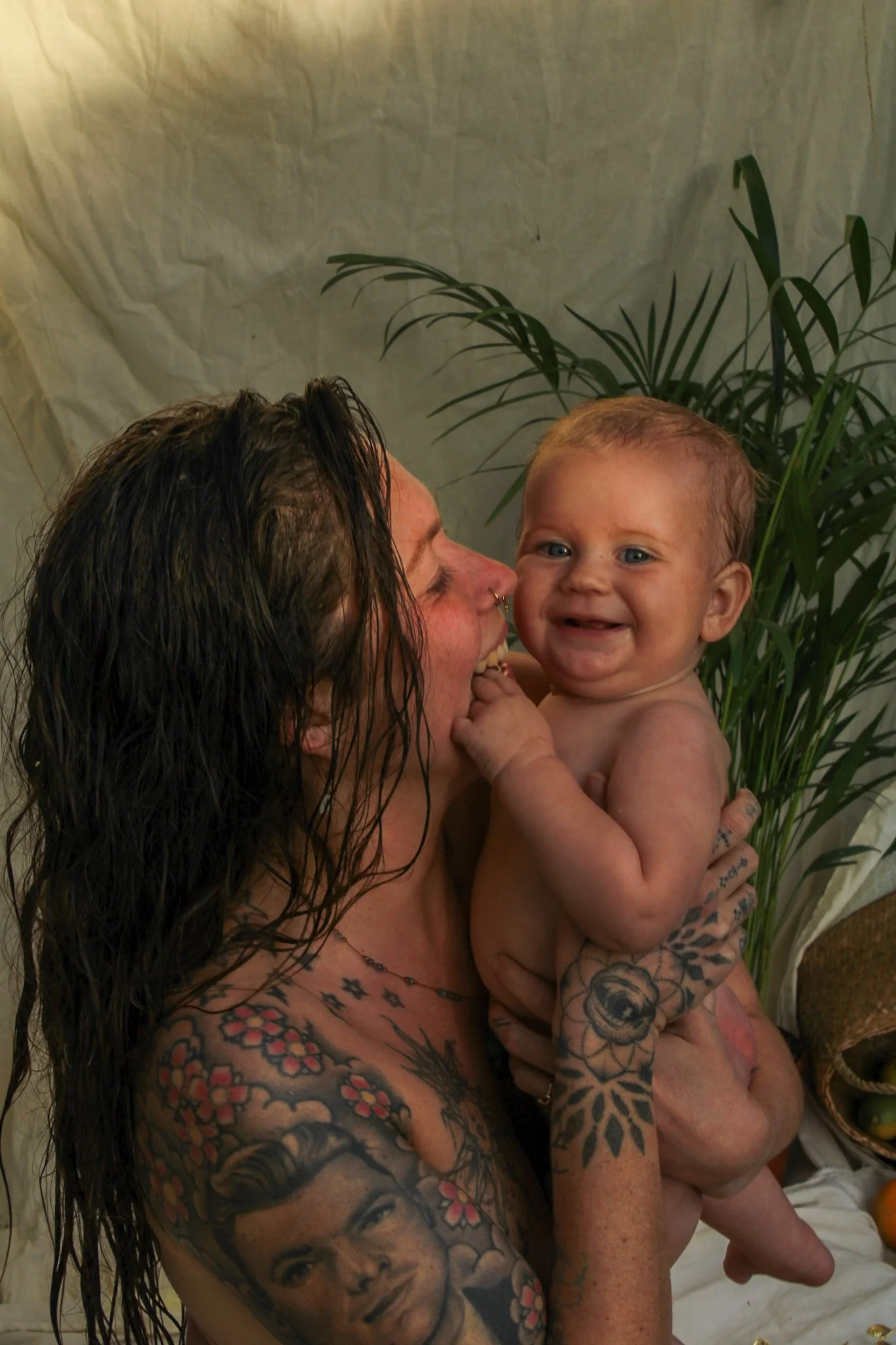 A woman with tattoos holding a smiling baby. The woman has dark, wet hair and facial piercings, and large tattoos on her arms and chest. The baby has light hair, blue eyes, and is smiling with a gap-toothed grin.