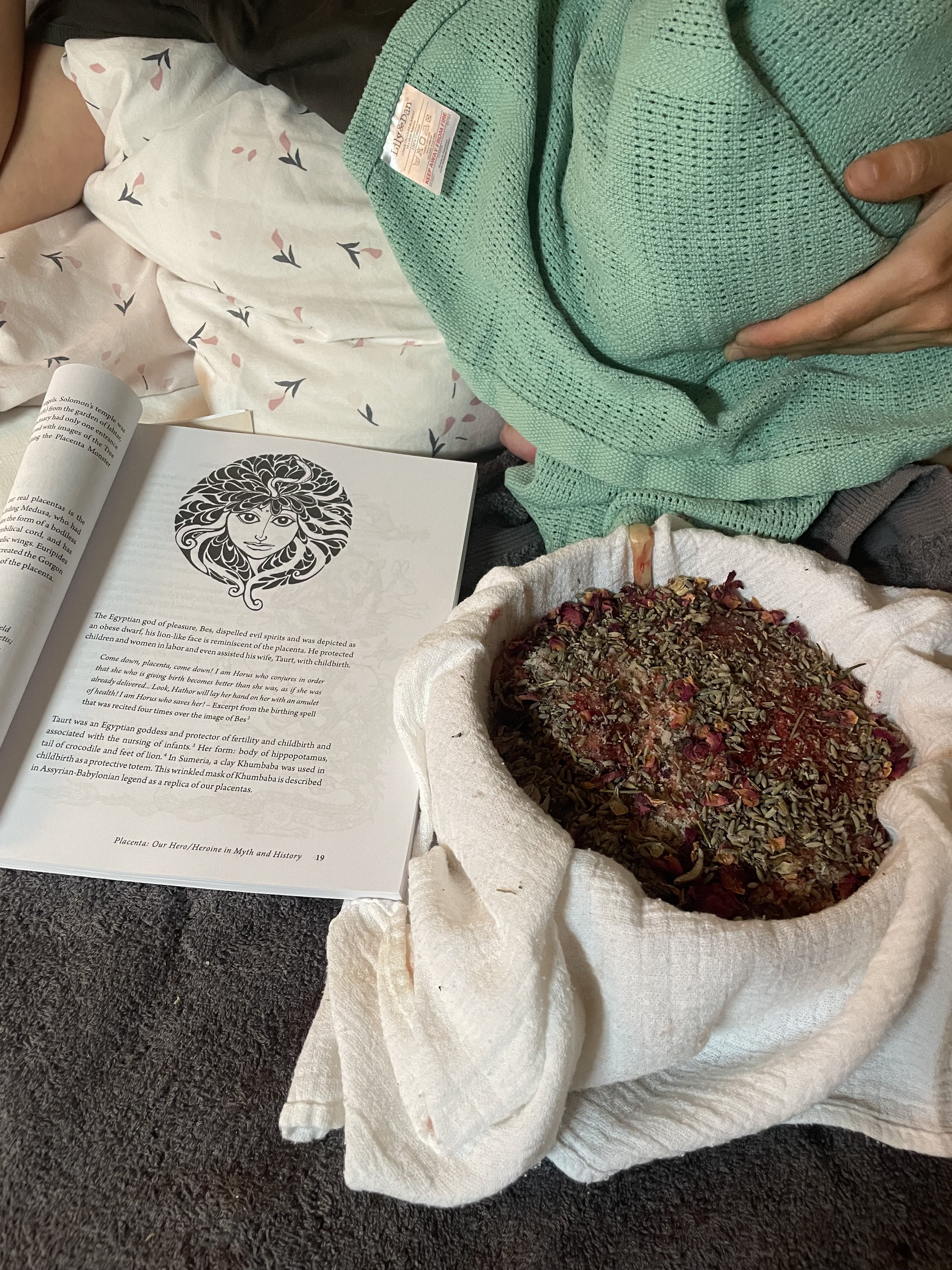 Close-up of a person wearing a green sweater, lying on a bed with white and pink bedsheets, reading a book about Egyptian mythology, with a mortar filled with herbs and dried flowers nearby.