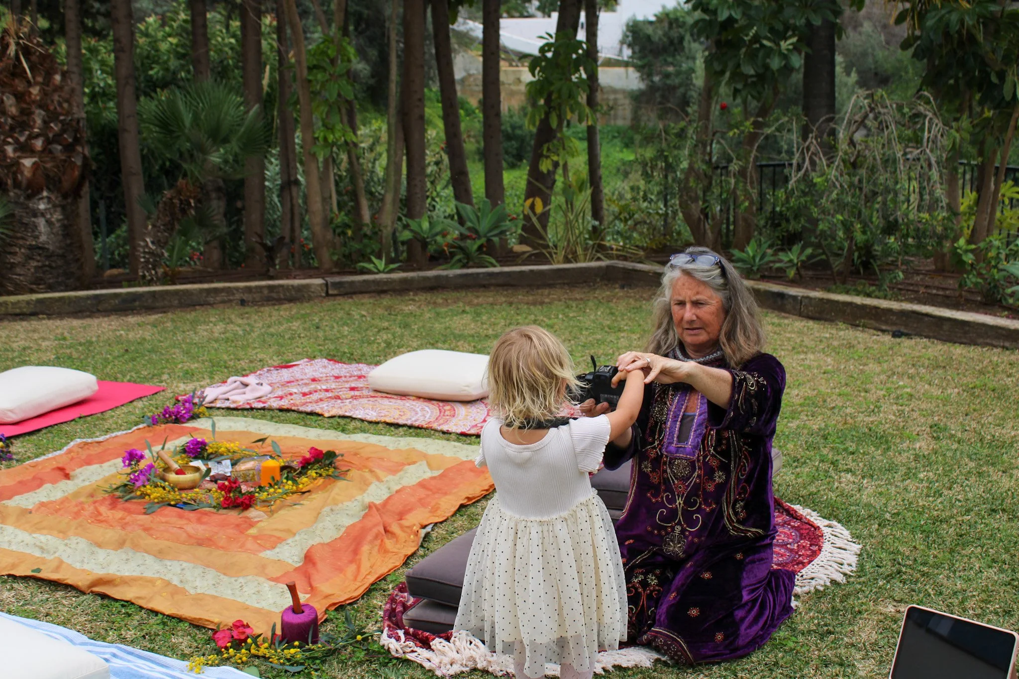 An elderly woman and a young girl in a garden, with the woman taking a photo of the girl. There are colorful fabric mats, pillows, and flowers on the grass, suggesting a spiritual or ceremonial setting.