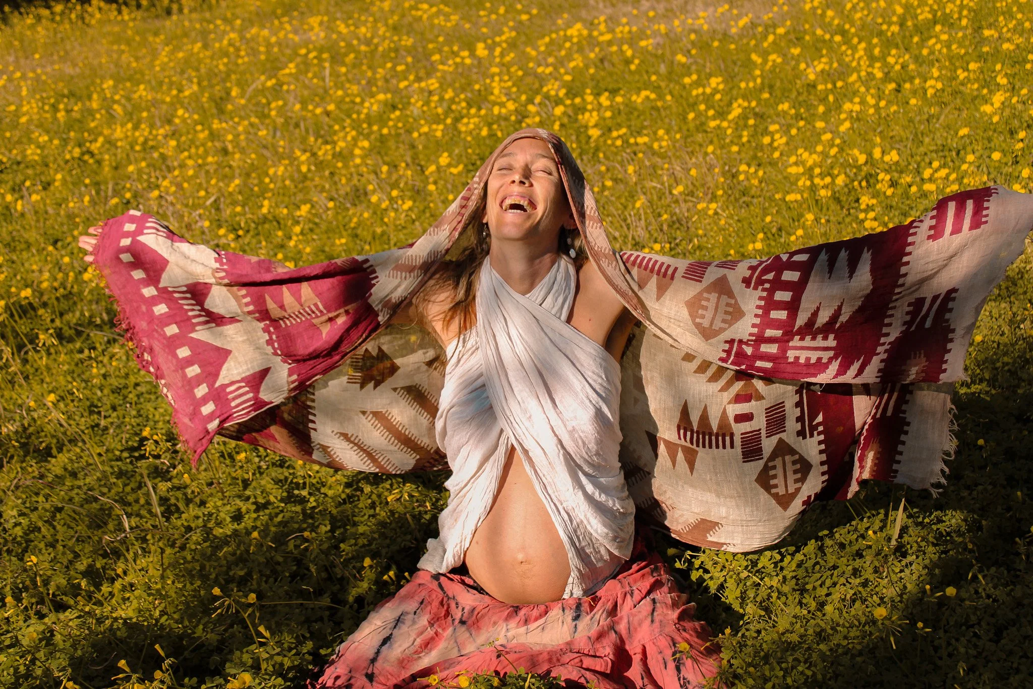 A woman celebrating outdoors in a field of yellow flowers, wearing a white dress and a patterned shawl, with arms outstretched and a joyful expression.
