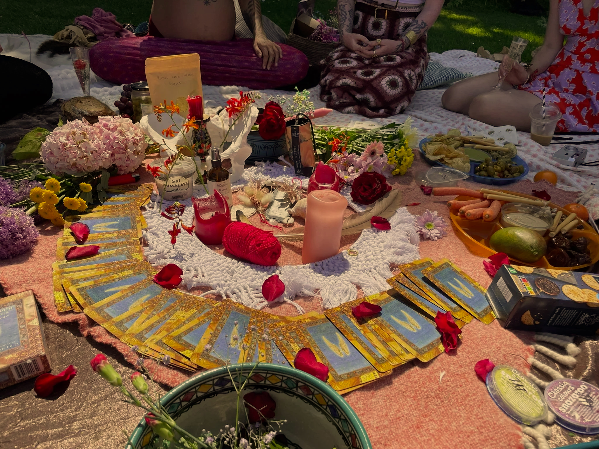 A colorful outdoor scene with a table covered in tarot cards, flowers, candles, and various items for a spiritual or ritualistic gathering, with people sitting around it.