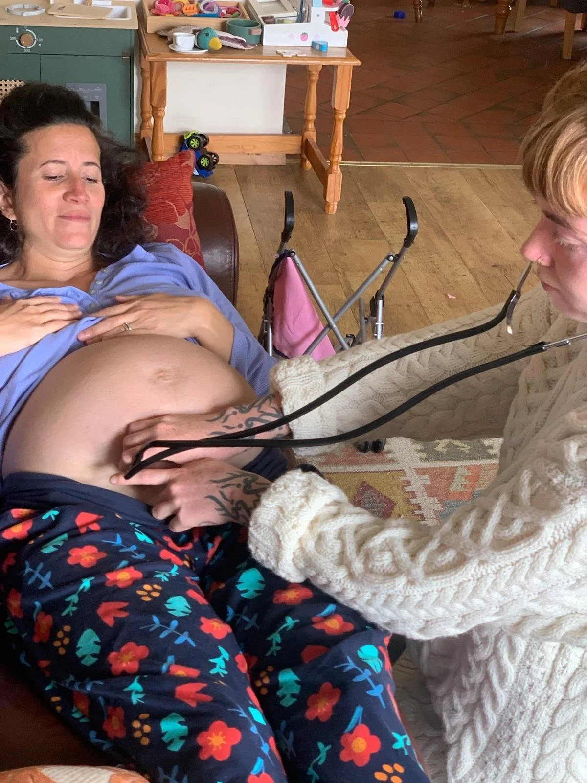 A woman sitting on a couch with her hand on her pregnant belly, while another woman in a white sweater with gloves checks her belly with a stethoscope. The woman on the couch has dark hair and is wearing a blue shirt and navy pants with a colorful fl